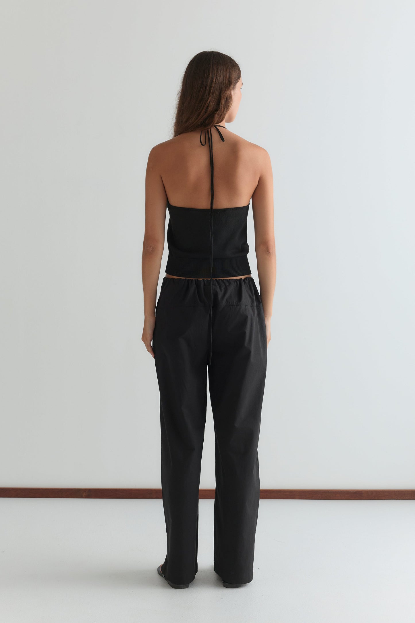 A woman faces away from the camera, wearing the Drawcord Knit Top - Black by Deiji Studios with an adjustable ruched neckline and loose black pants, set against a plain white background.
