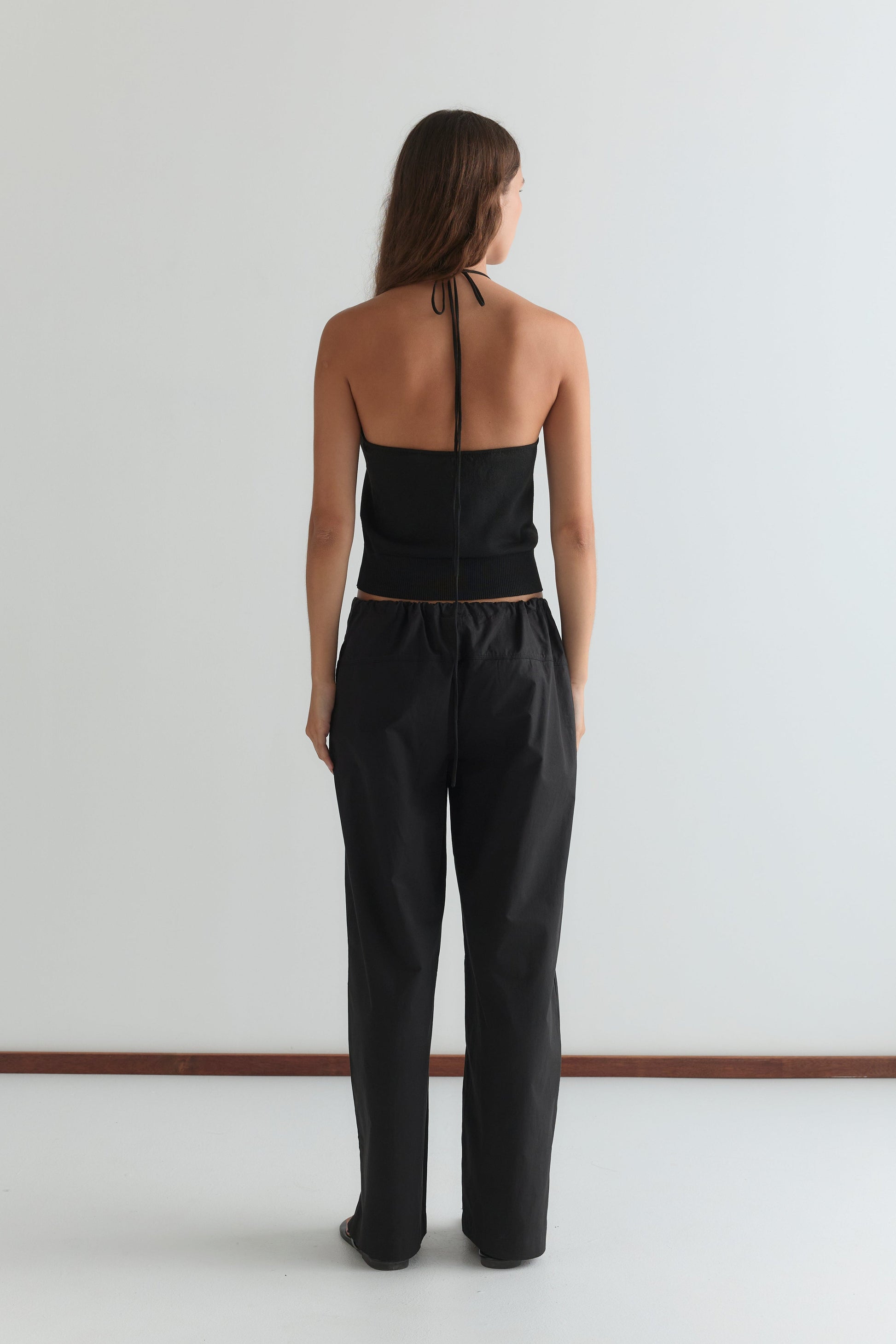 A woman faces away from the camera, wearing the Drawcord Knit Top - Black by Deiji Studios with an adjustable ruched neckline and loose black pants, set against a plain white background.