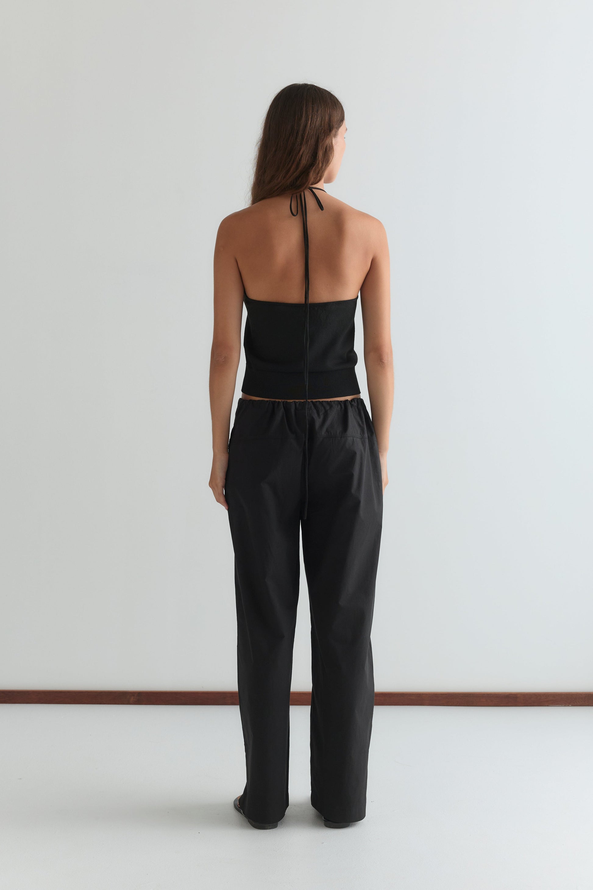 A person with long hair, facing away, wears the Seam Pant - Black and a black halter-top, standing against a plain white wall and floor.