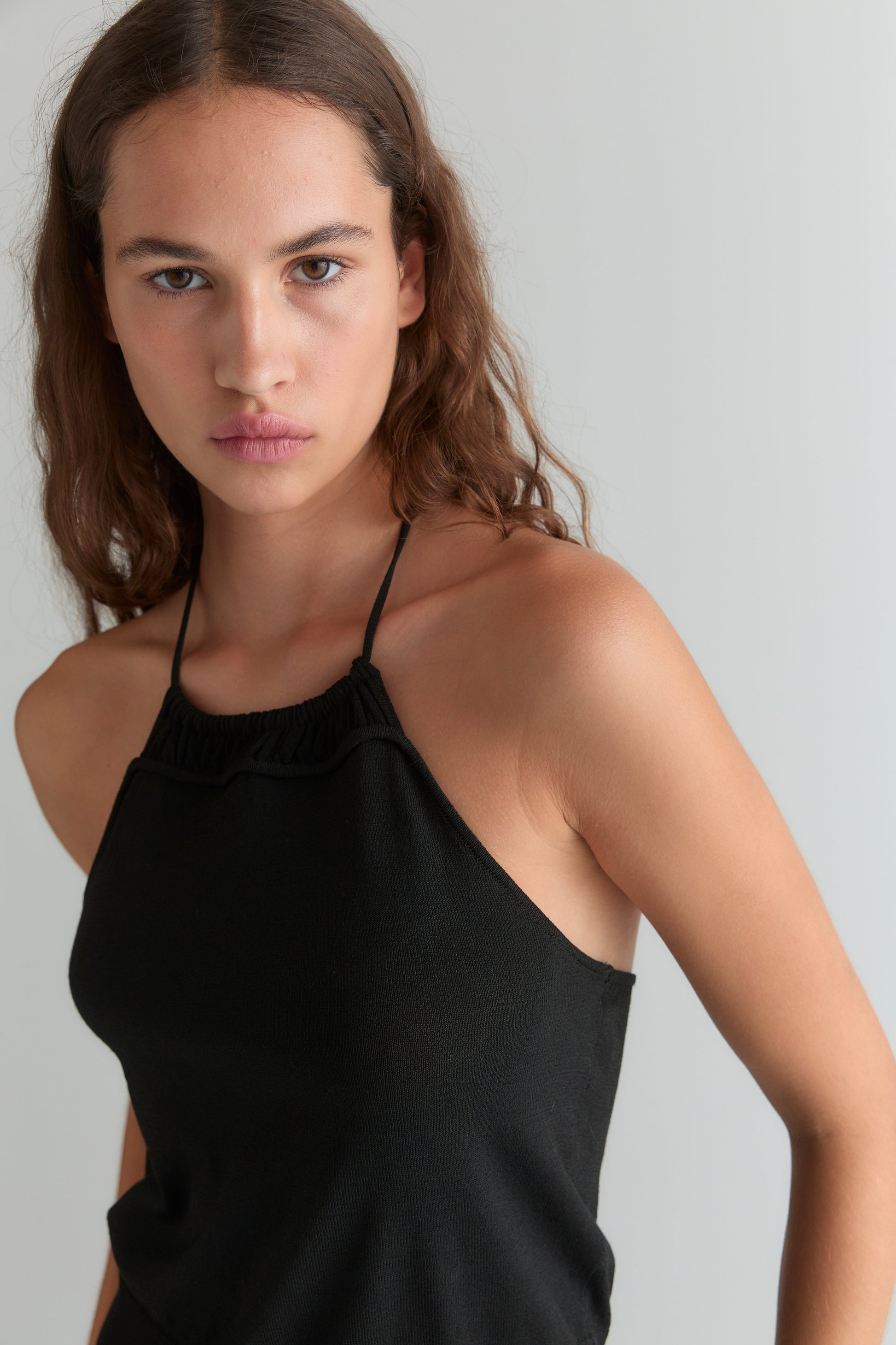 A young woman with long brown hair wears the Drawcord Knit Top - Black by Deiji Studios, featuring an adjustable ruched neckline. She stands against a plain light gray background, gazing seriously at the camera.