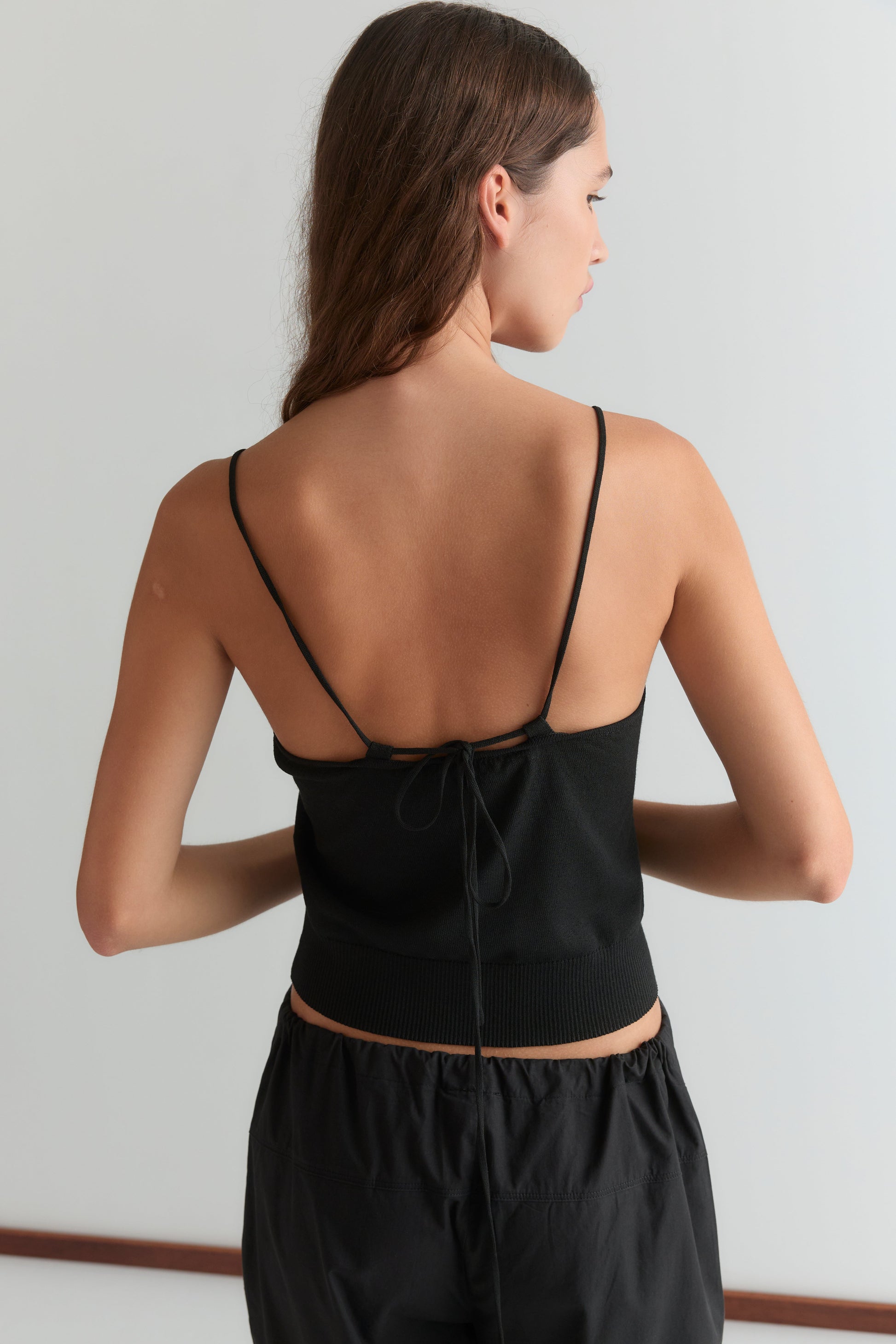 A woman with long brown hair faces away, wearing the Deiji Studios Drawcord Knit Top - Black with an adjustable ruched neckline and open back, paired with black pants against a plain light background.