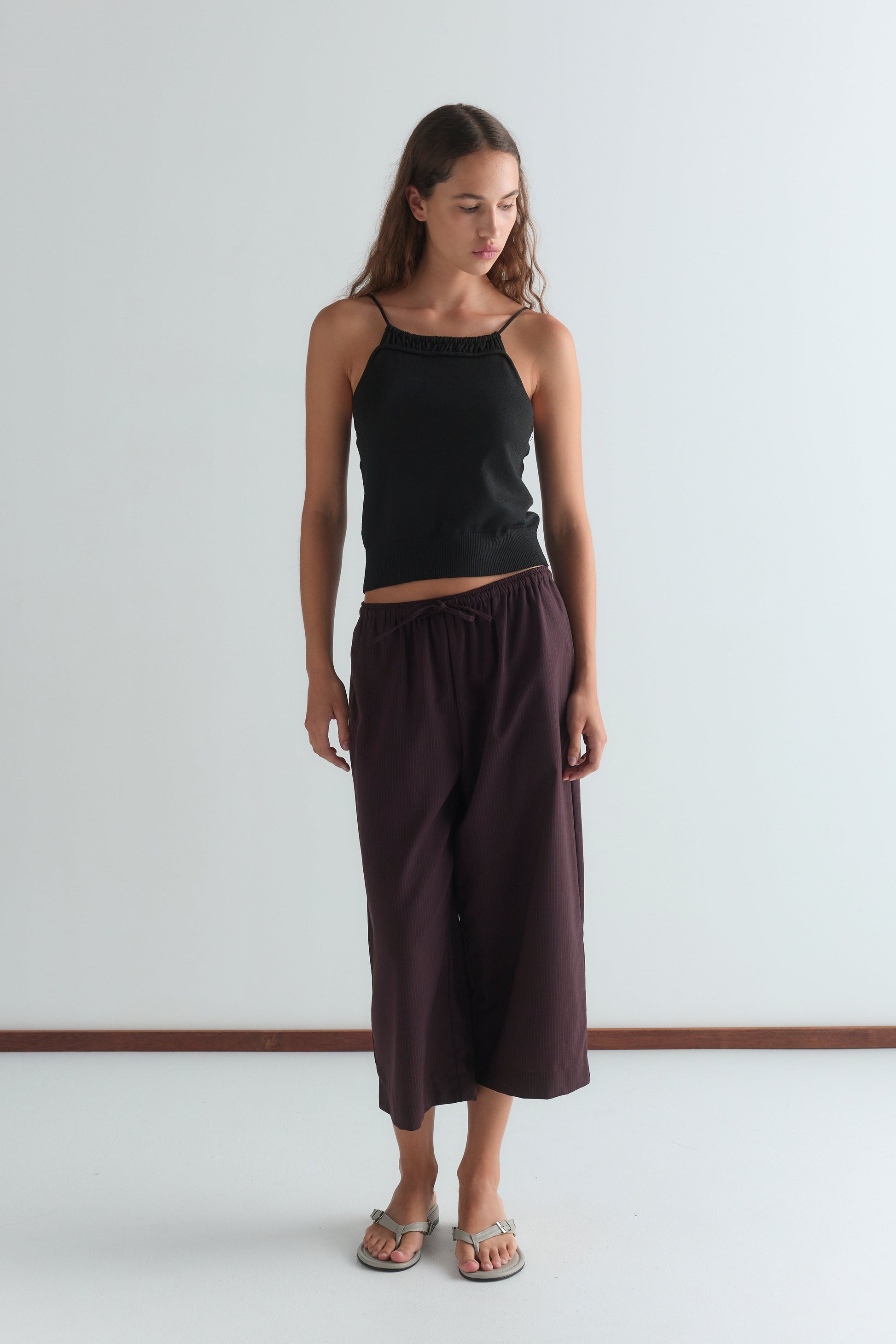 A woman stands indoors against a plain white wall, wearing a Deiji Studios black sleeveless top, Drawcord Culotte - Burgundy Brown Stripe pants, and sandals. She looks down with her arms relaxed at her sides.