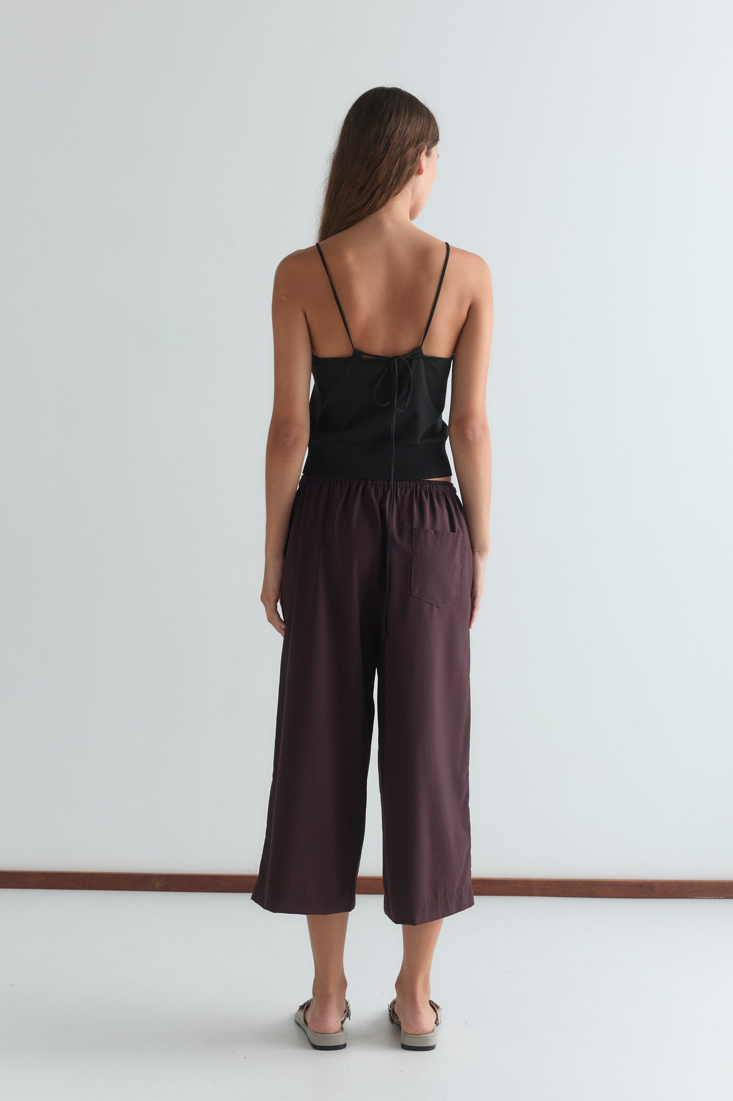 A woman with long brown hair faces away from the camera, wearing a relaxed black spaghetti strap top and Drawcord Culotte - Burgundy Brown Stripe pants in a minimalist room with a white wall and brown floor trim.