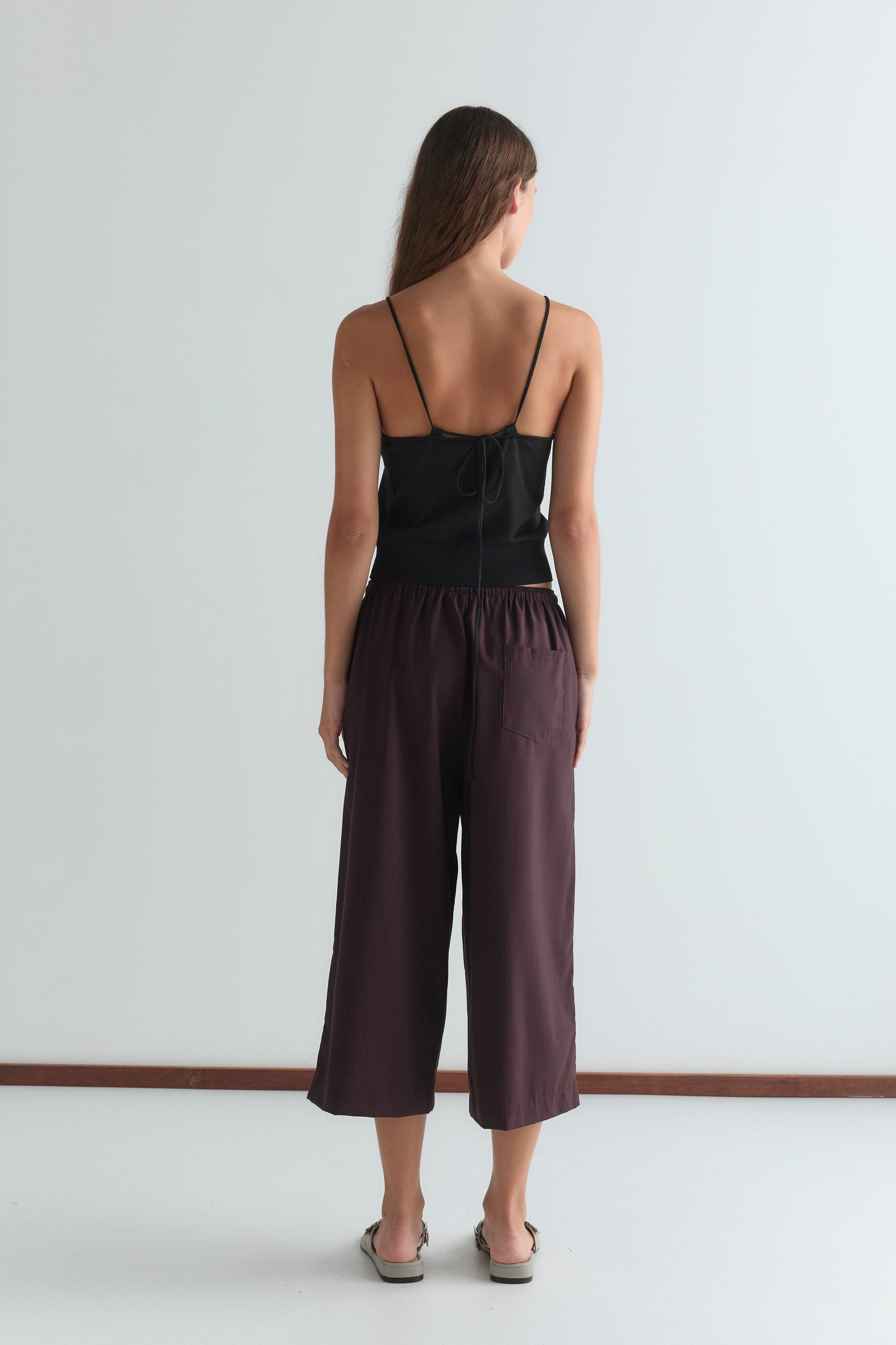 A woman with long brown hair faces away from the camera, wearing a relaxed black spaghetti strap top and Drawcord Culotte - Burgundy Brown Stripe pants in a minimalist room with a white wall and brown floor trim.