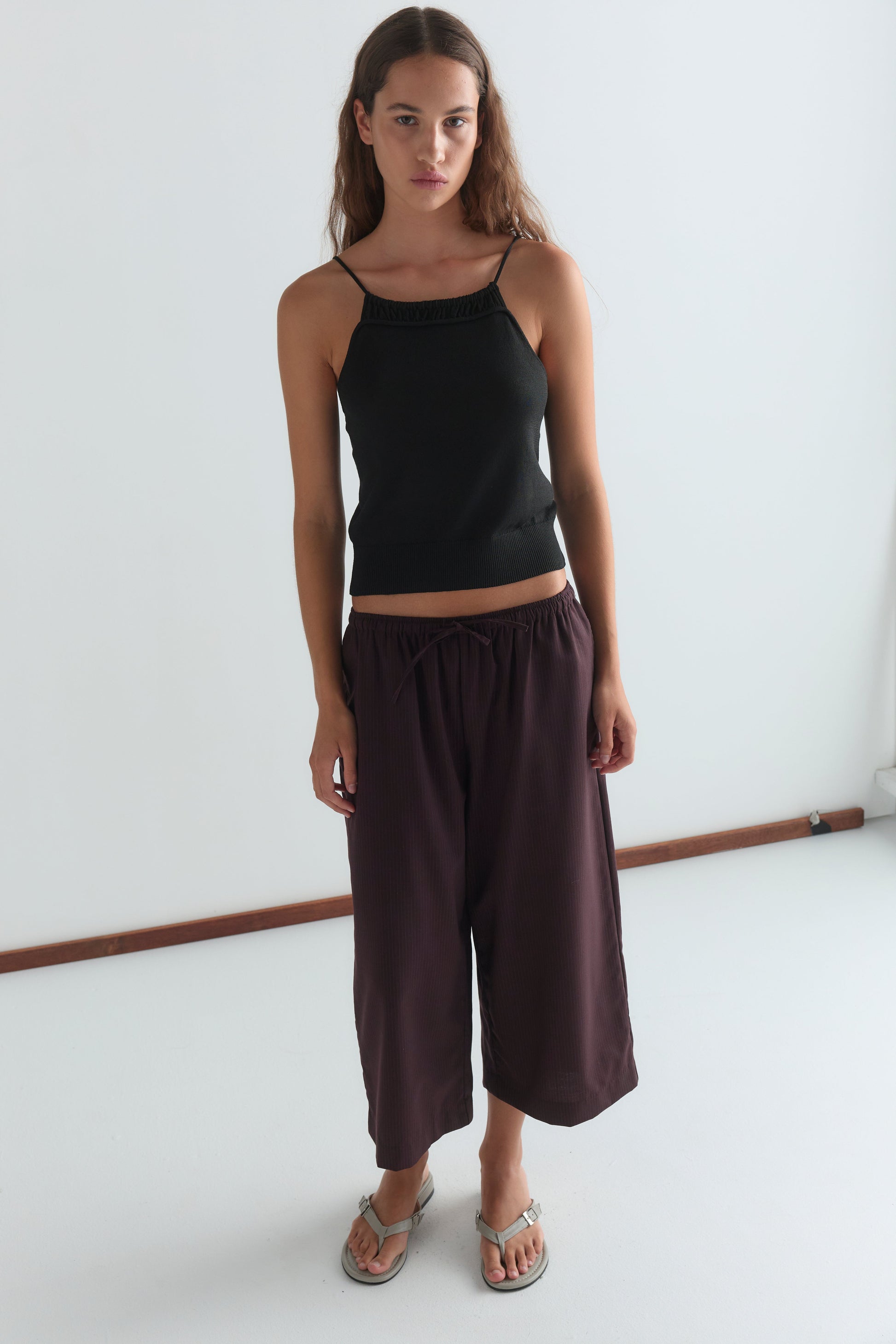 A woman with long brown hair wears a black sleeveless top, Drawcord Culotte - Burgundy Brown Stripe by Deiji Studios, and sandals, standing against a plain white background.