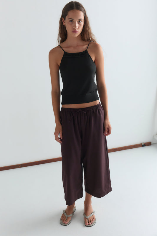 A woman with long brown hair wears a black sleeveless top, Drawcord Culotte - Burgundy Brown Stripe by Deiji Studios, and sandals, standing against a plain white background.