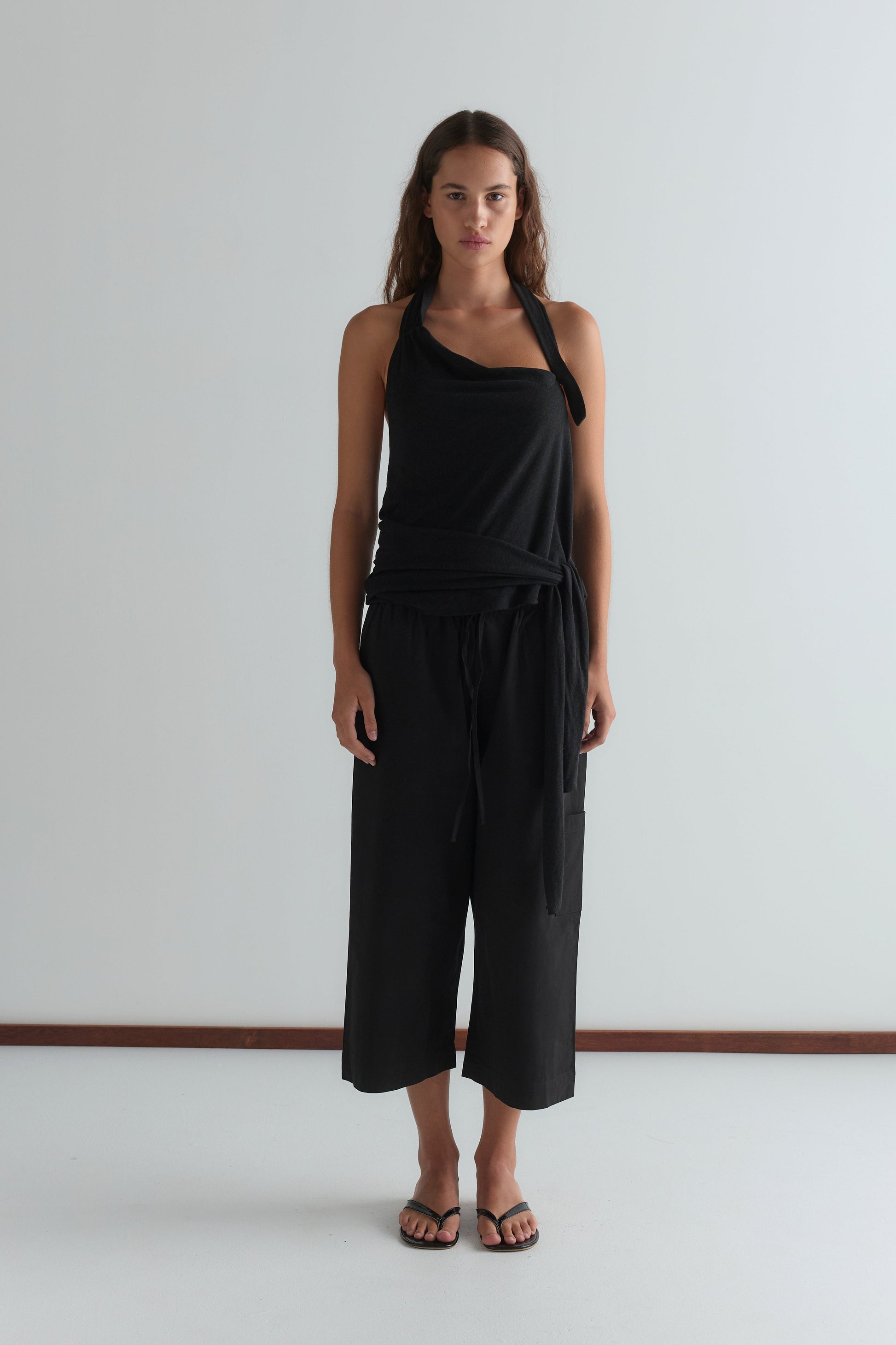 A woman with long, wavy brown hair and a neutral expression stands against a plain white wall wearing a sleeveless black halter top, Mid Pocket Short - Black by Deiji Studios, and black flip-flops.