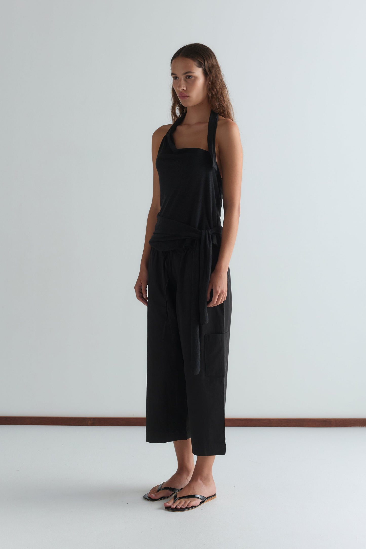 A woman with long brown hair wears a sleeveless black top and Deiji Studios Mid Pocket Short - Black, paired with black flip-flops. She stands against a plain white wall, looking slightly to the side.