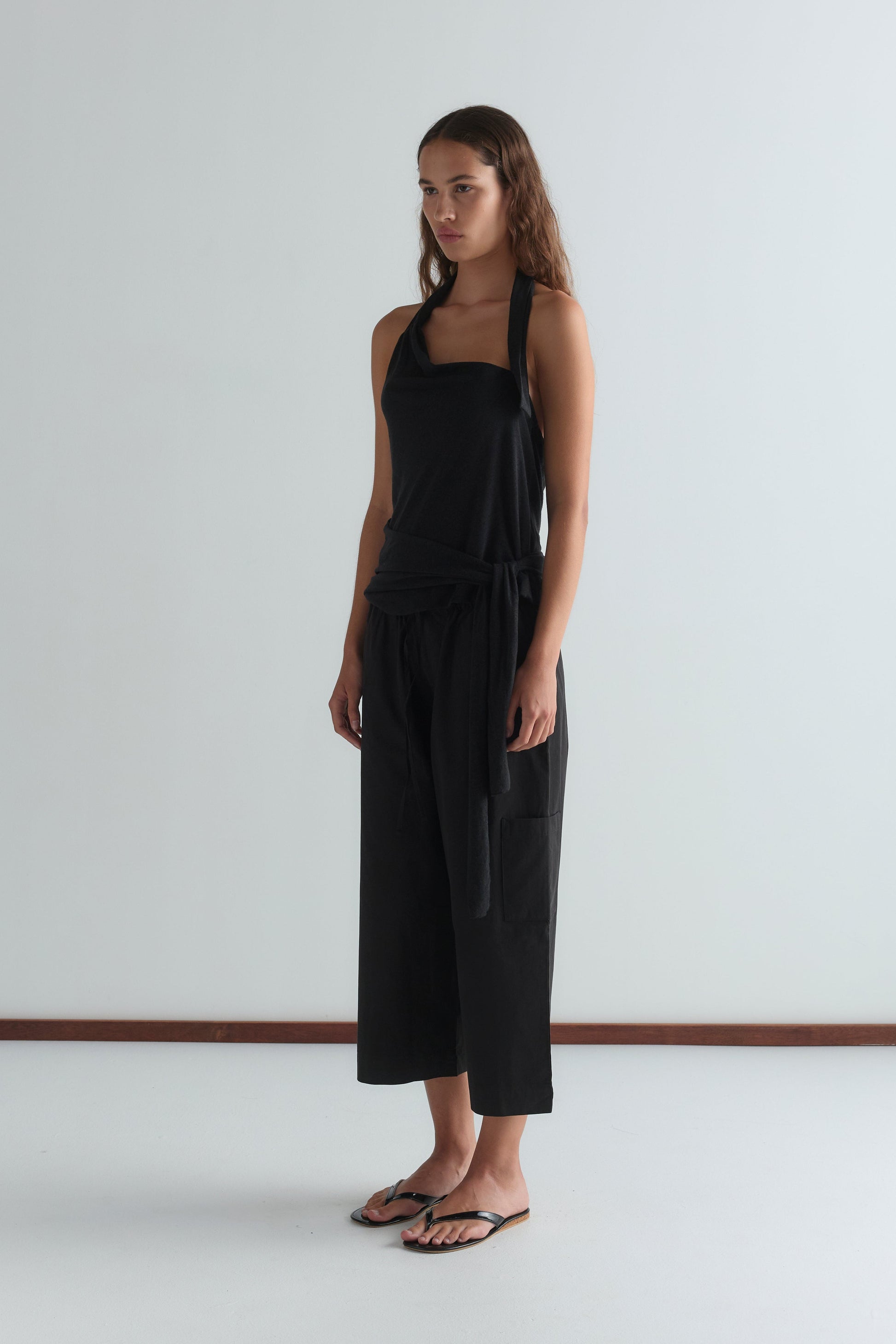 A woman with long brown hair wears a sleeveless black top and Deiji Studios Mid Pocket Short - Black, paired with black flip-flops. She stands against a plain white wall, looking slightly to the side.