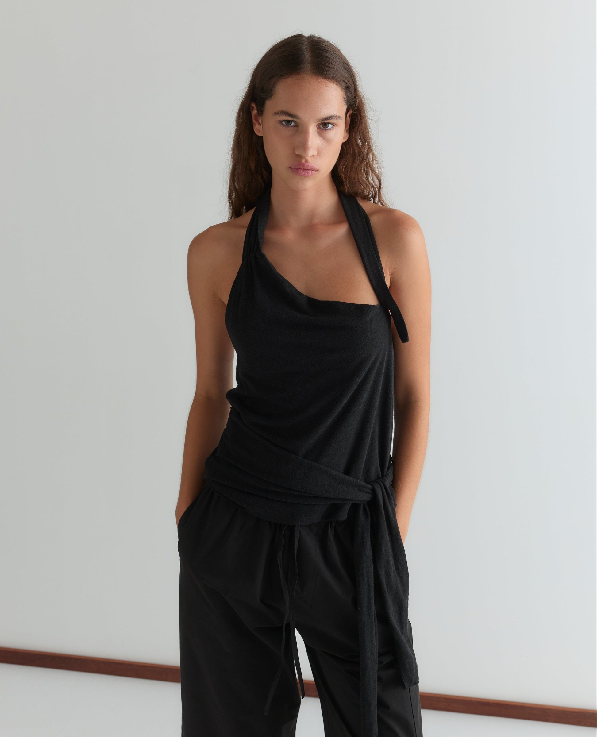 A woman with long brown hair stands against a white wall, wearing the Deiji Studios Drape Halter in Black and black pants, hands in her pockets, looking directly at the camera with a neutral expression.