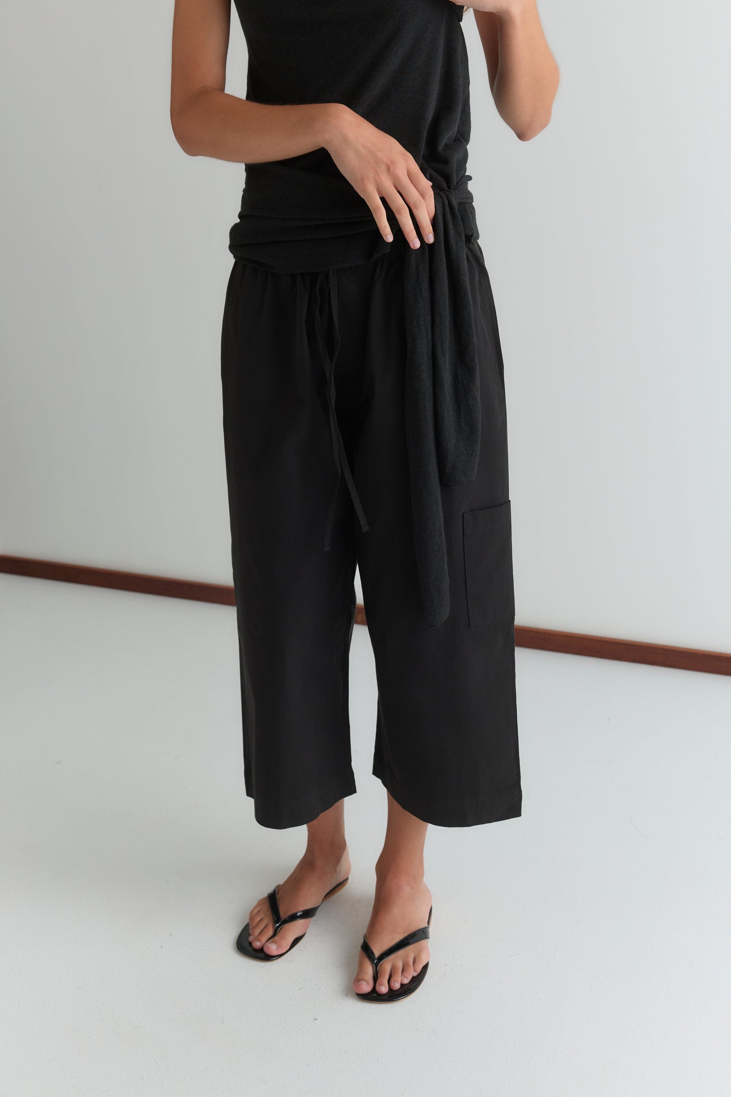 A person wears a Deiji Studios black sleeveless top with the Mid Pocket Short - Black, styled as relaxed 3/4 wide-leg cropped pants, and black flip-flops, standing on a white floor against a plain light wall with their face not visible.
