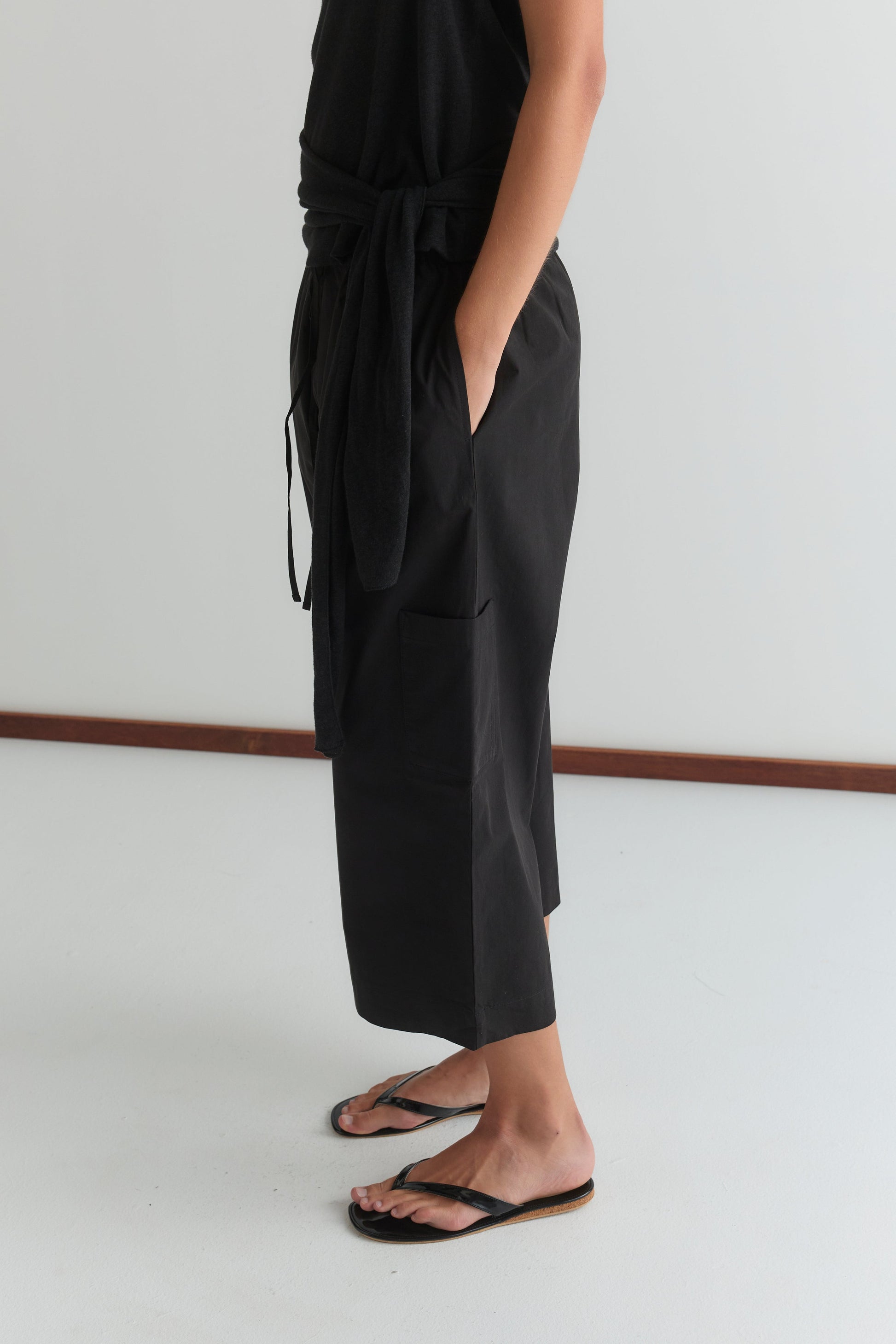 A person stands indoors on a white floor, wearing a black shirt tied at the waist and the Mid Pocket Short - Black from Deiji Studios, paired with black flip-flops. Their hands are tucked into the shorts' pockets.
