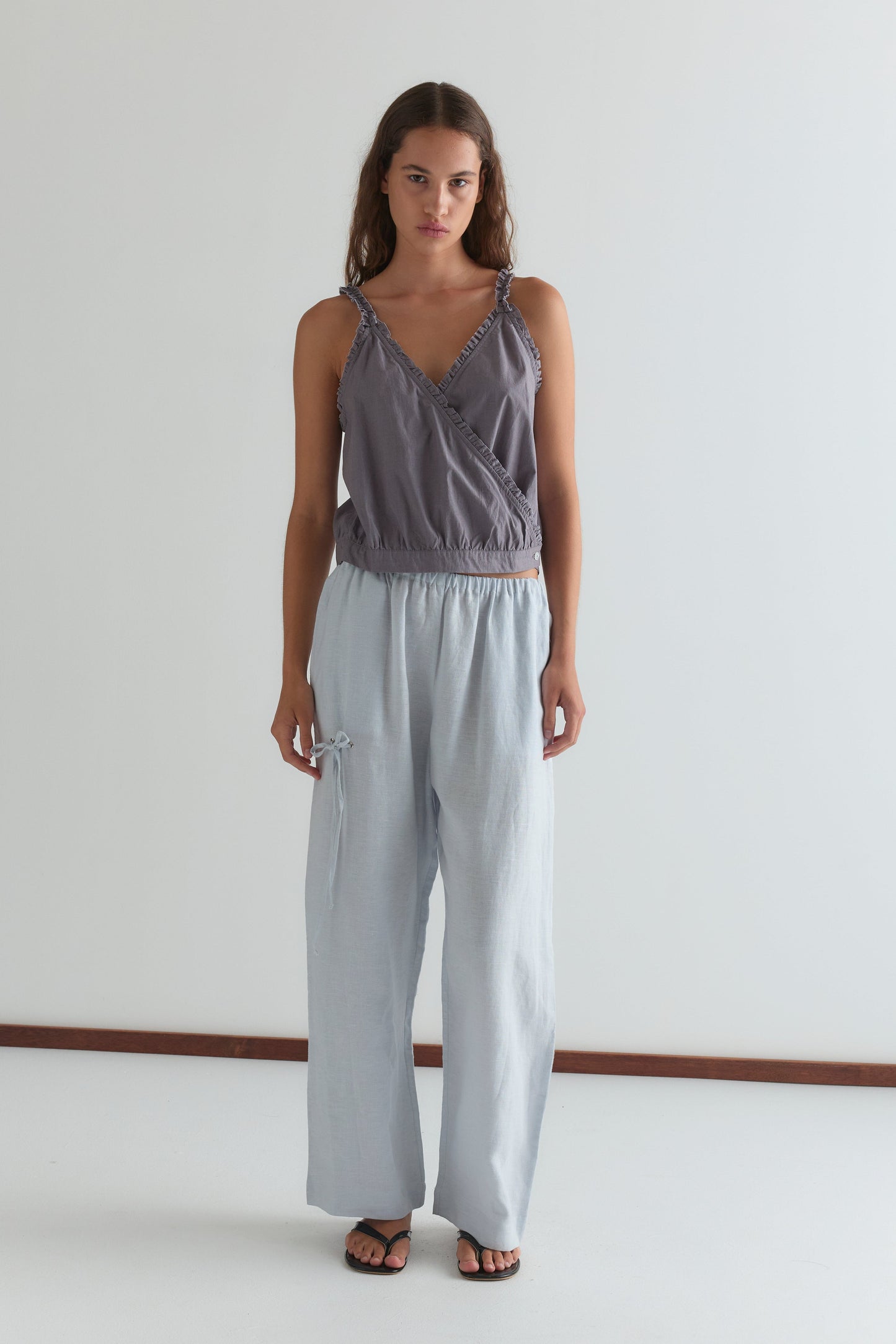A woman with long brown hair and a neutral expression stands against a white wall, wearing Deiji Studios’ sleeveless gray wrap top, Eyelet Pant in Soft Blue, and black sandals.