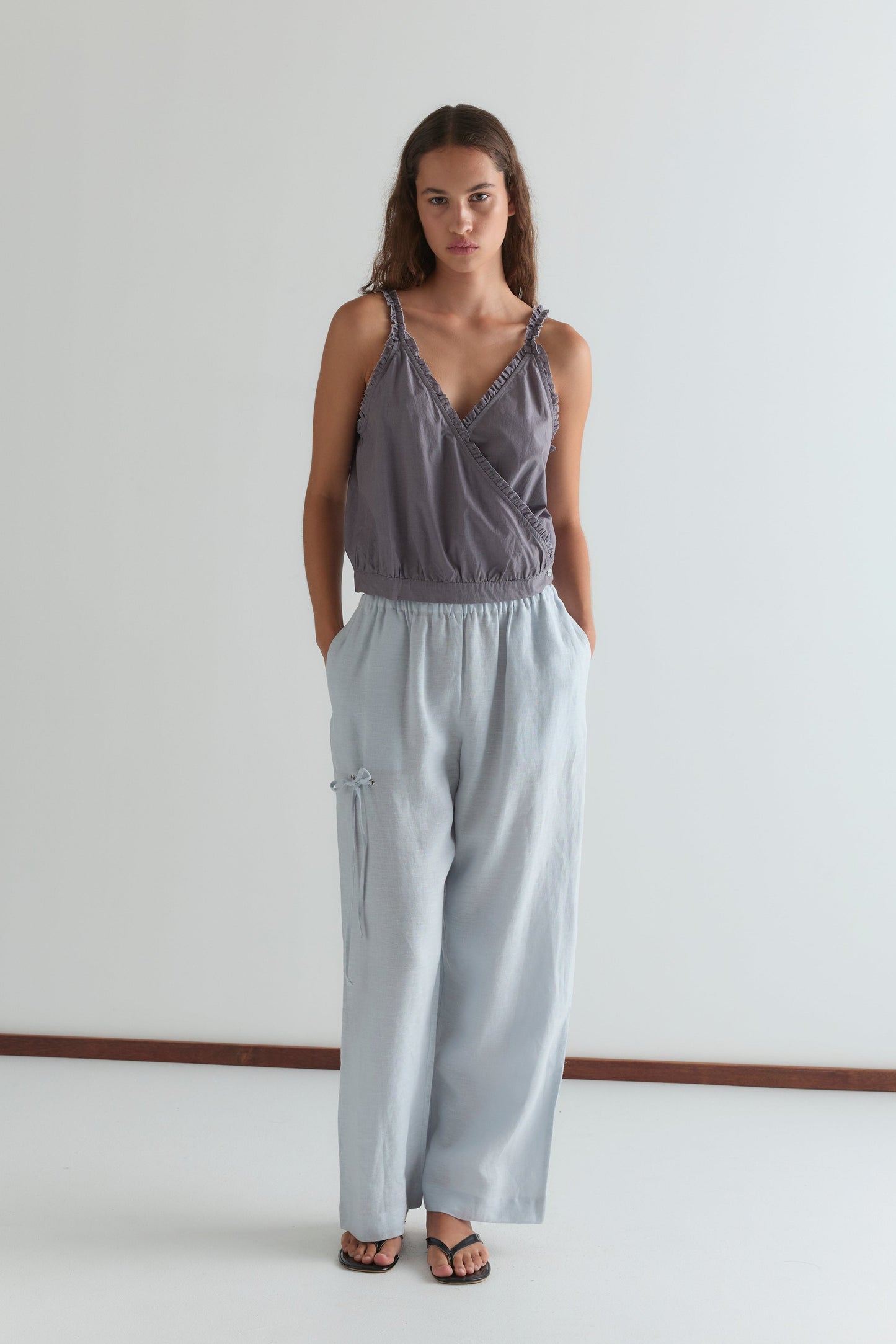 A woman stands against a plain white wall wearing Deiji Studios’ loose, sleeveless Overlie Top in Currant and light blue wide-leg pants with her hands in her pockets. She has long brown hair and wears black sandals.