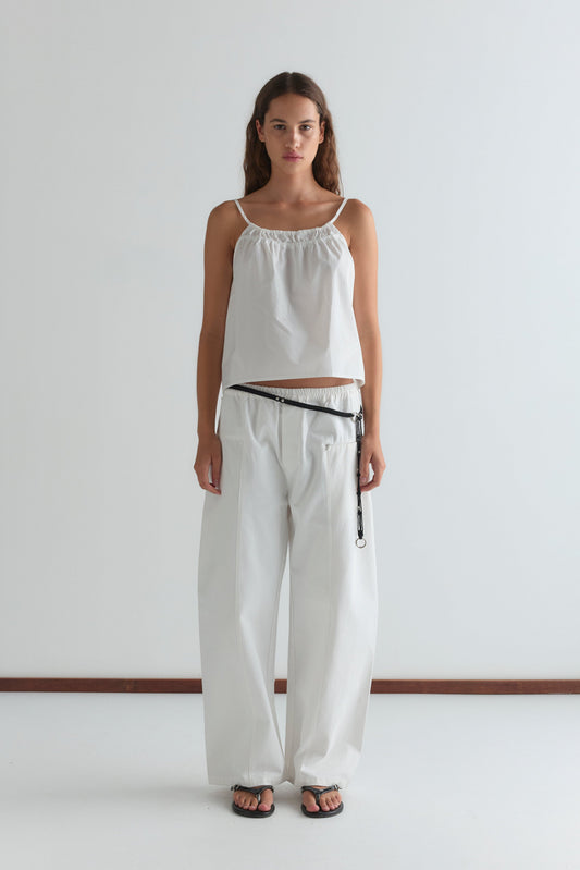 A woman with long brown hair stands against a white wall, wearing the Pleat Pant - White with a loose white sleeveless top, black sandals, and a black belt. She faces the camera neutrally.