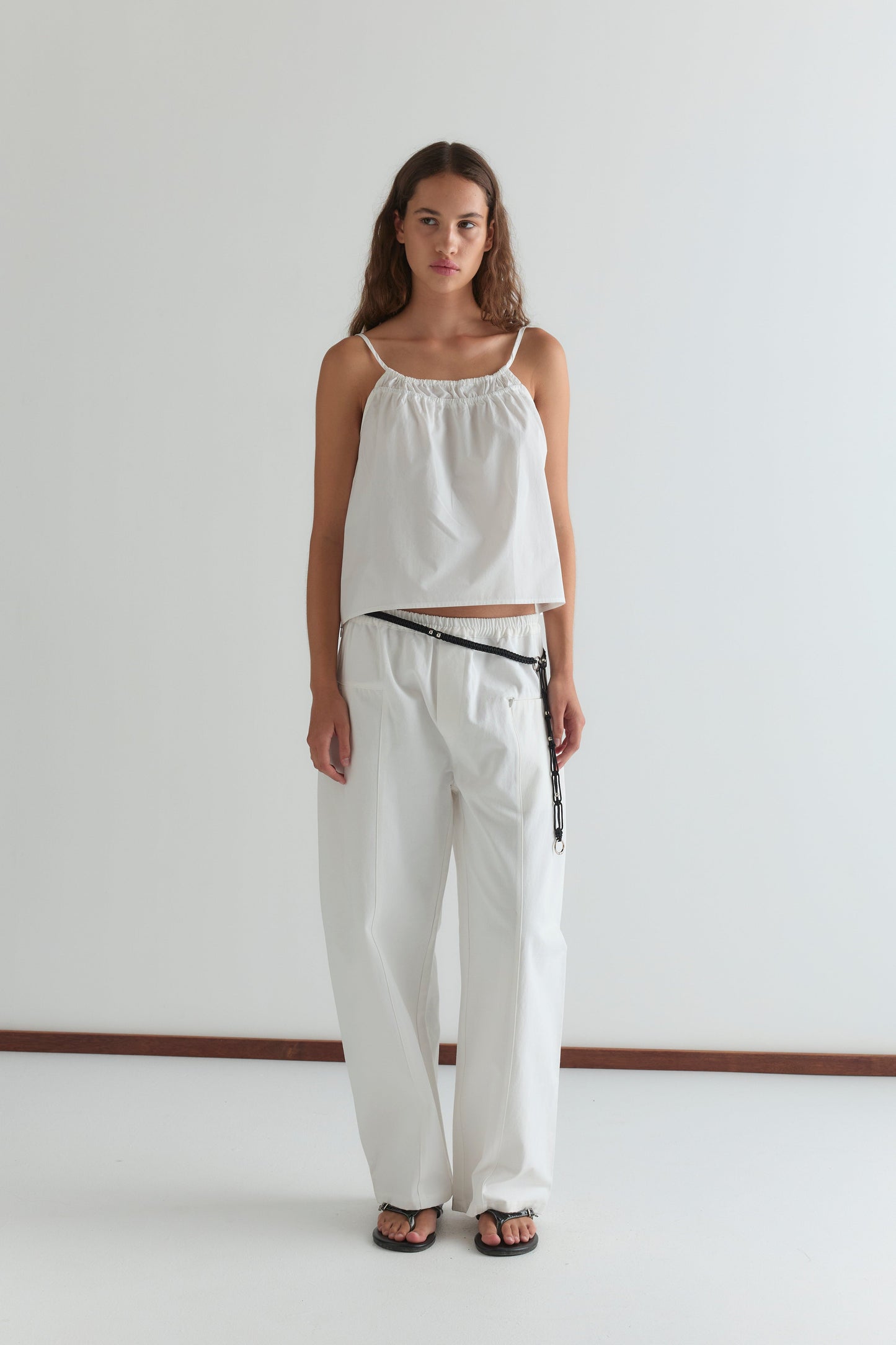 A woman with long brown hair and a neutral expression stands against a plain white wall, wearing the Pleat Pant - White paired with a loose white sleeveless top, black belt, and black sandals.