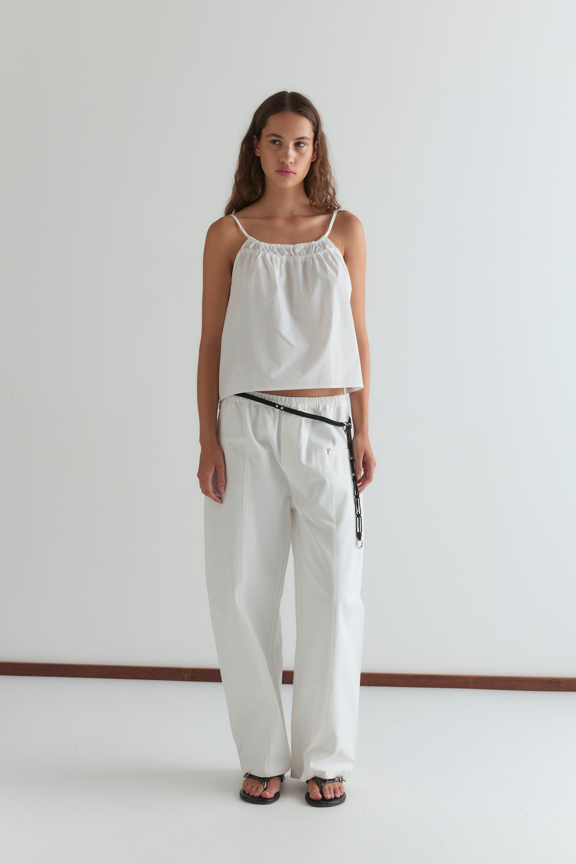 A woman with long brown hair and a neutral expression stands against a plain white wall, wearing the Pleat Pant - White paired with a loose white sleeveless top, black belt, and black sandals.