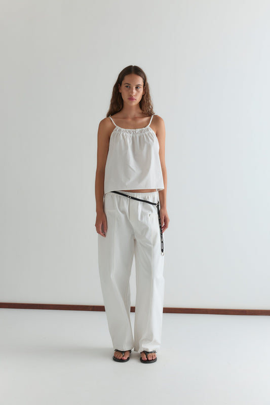 A person with long brown hair and a neutral expression stands against a plain white wall wearing a sleeveless white top, the Pleat Pant - White from Deiji Studios, a black belt, and black sandals.