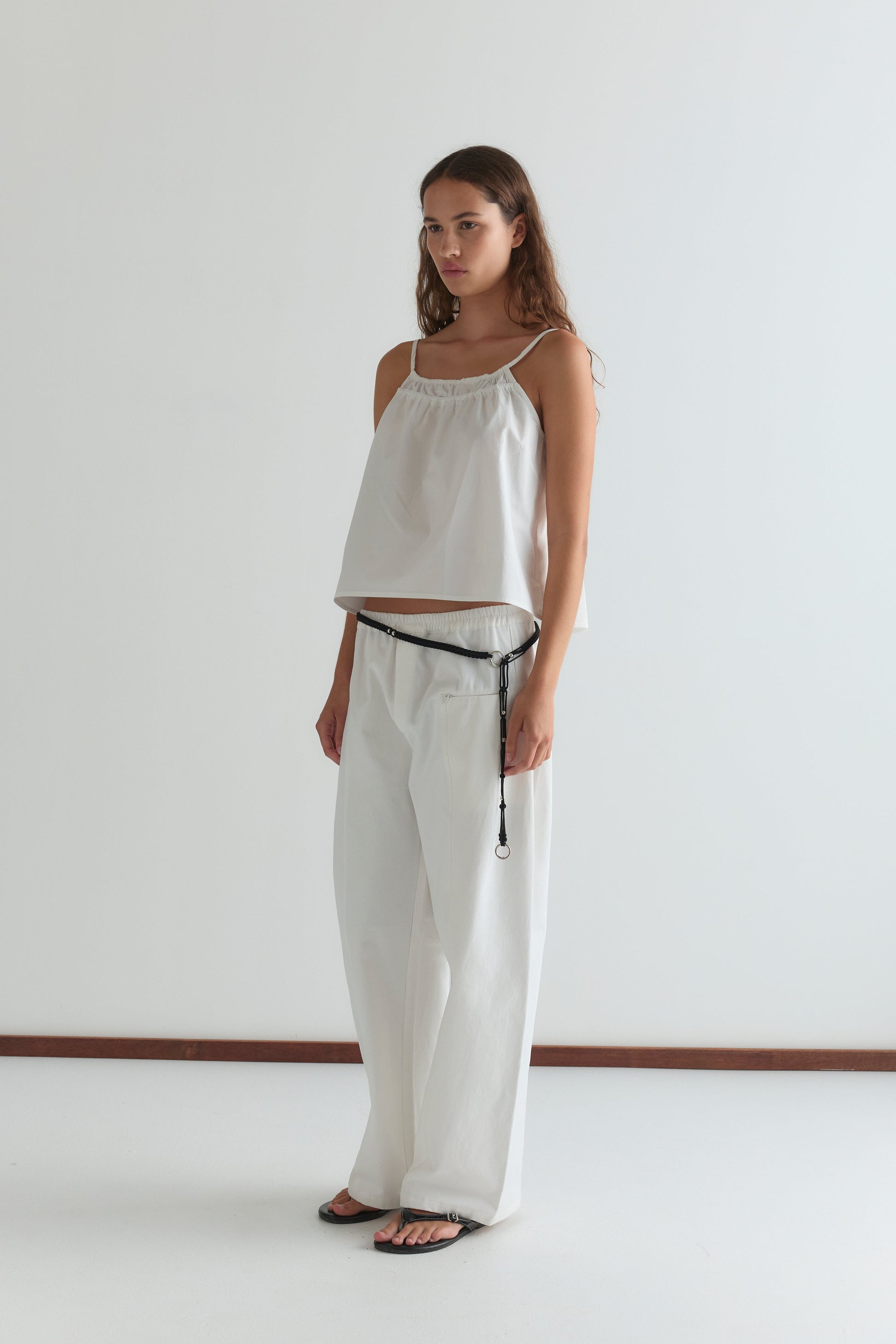 A woman with long wavy hair and a neutral expression stands against a plain white background wearing the white Pleat Pant, a loose sleeveless top, black sandals, and a thin black belt with a loop.