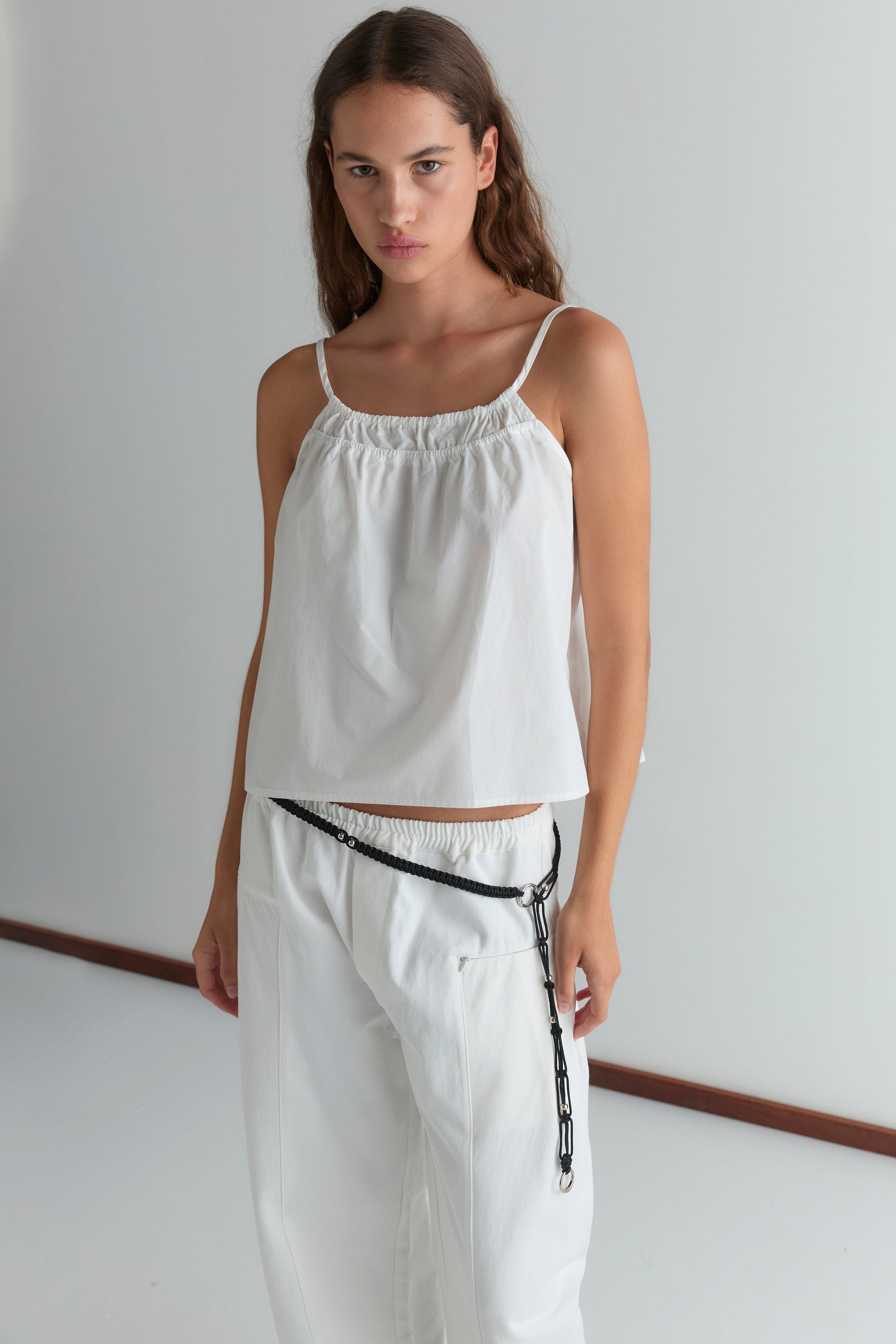 A woman wearing loose white Pleat Pants and a sleeveless top with a black chain belt stands against a plain white background, gazing confidently at the camera.