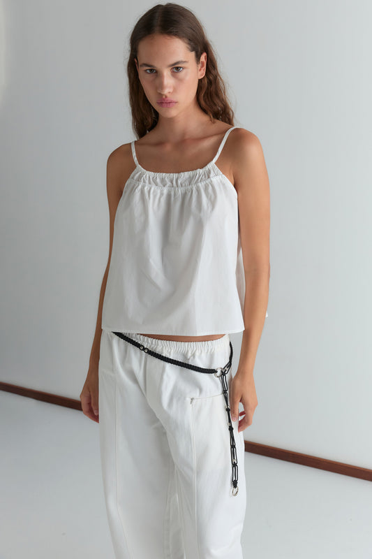 A woman with long brown hair stands indoors wearing the Pleat Pant - White, featuring a relaxed straight leg and elasticated waistband. She pairs it with a sleeveless white top and a black chain accessory. The background is plain and minimalistic.