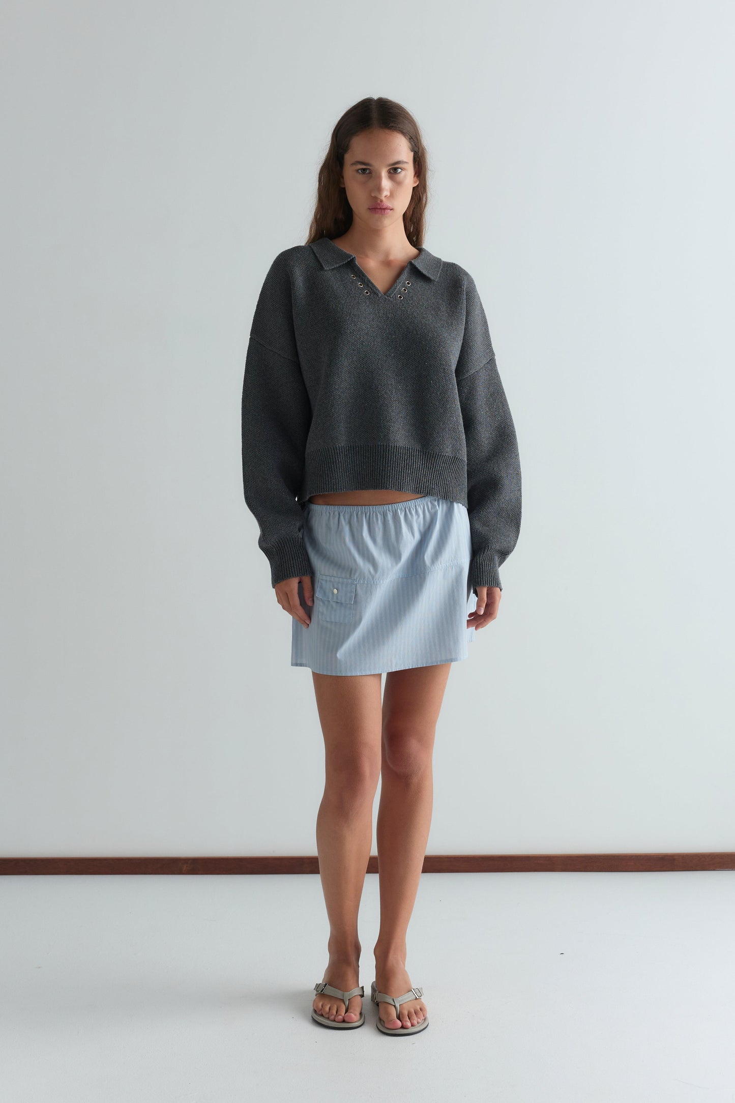 A woman with long brown hair and a neutral expression stands against a white wall wearing Deiji Studios' Eyelet Knit - Dark Pebble sweater, a light blue pocketed mini skirt, and beige sandals.