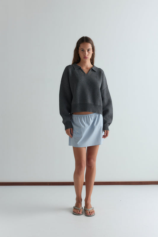 A woman with long brown hair stands indoors against a white wall, wearing the Deiji Studios Eyelet Knit - Dark Pebble sweater, a light blue mini skirt, and sandals. She has a neutral expression.