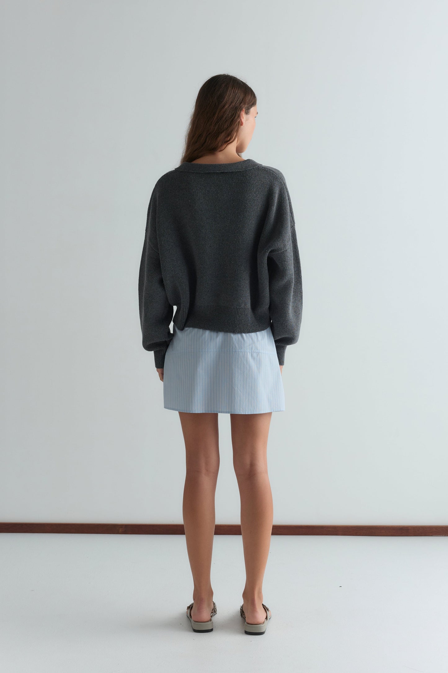 A woman faces away from the camera, wearing the Eyelet Knit - Dark Pebble by Deiji Studios, a light blue pinstriped mini skirt, and sandals in a minimal, light-toned room with a white floor and wall.