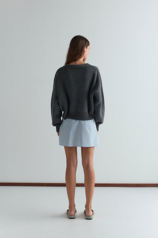A woman stands facing away from the camera, wearing Deiji Studios' Eyelet Knit - Dark Pebble sweater, a light blue mini skirt, and sandals in a minimalist room with a white floor and light-colored wall.