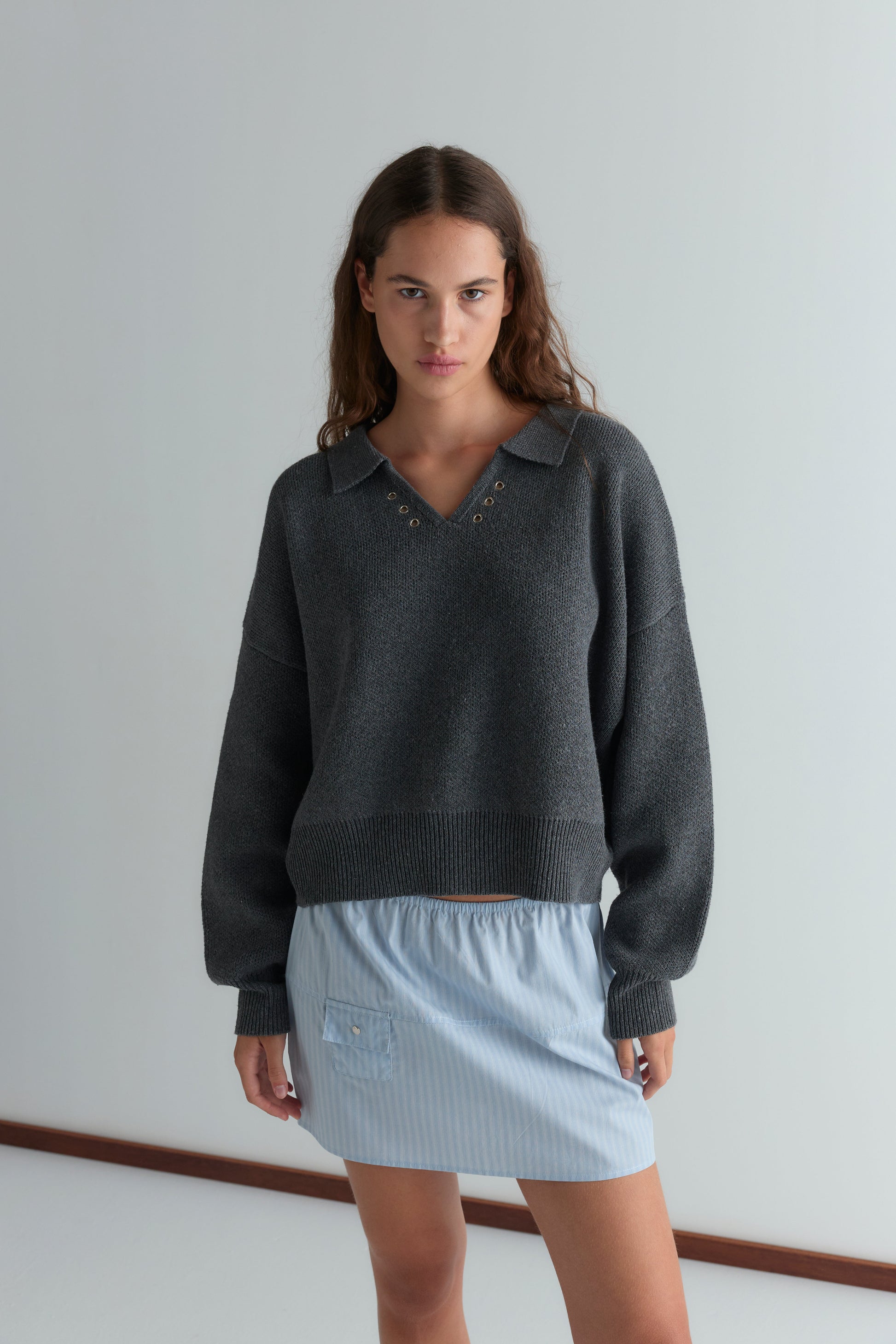 A woman with long brown hair wears the Eyelet Knit - Dark Pebble sweater from Deiji Studios, featuring grommet details at the collar, paired with a light blue striped mini skirt. She stands indoors by a white wall, gazing at the camera neutrally.