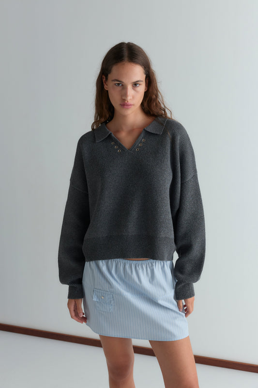 A young woman with wavy brown hair stands indoors, wearing the Eyelet Knit - Dark Pebble sweater paired with a light blue, short, pinstriped skirt. She looks at the camera with a neutral expression.