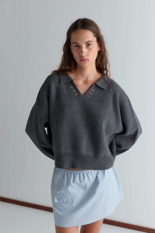 A woman with long brown hair wears the Eyelet Knit - Dark Pebble, a relaxed long-sleeve sweater with grommet details at the neckline, paired with a light blue mini skirt, standing against a plain light background.