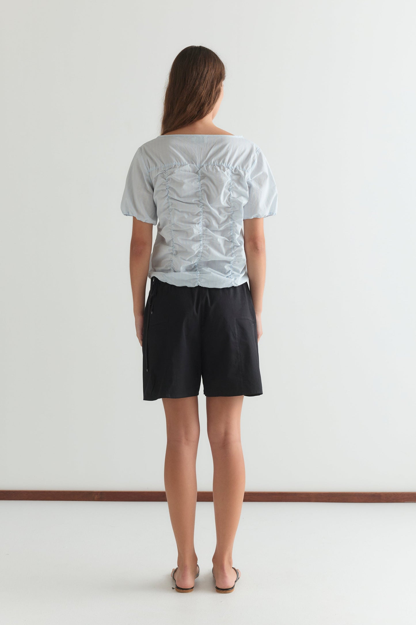 A woman with long brown hair, facing away, wears the Crinkle Top in Powder Blue Stripe with black shorts and sandals against a plain white background.