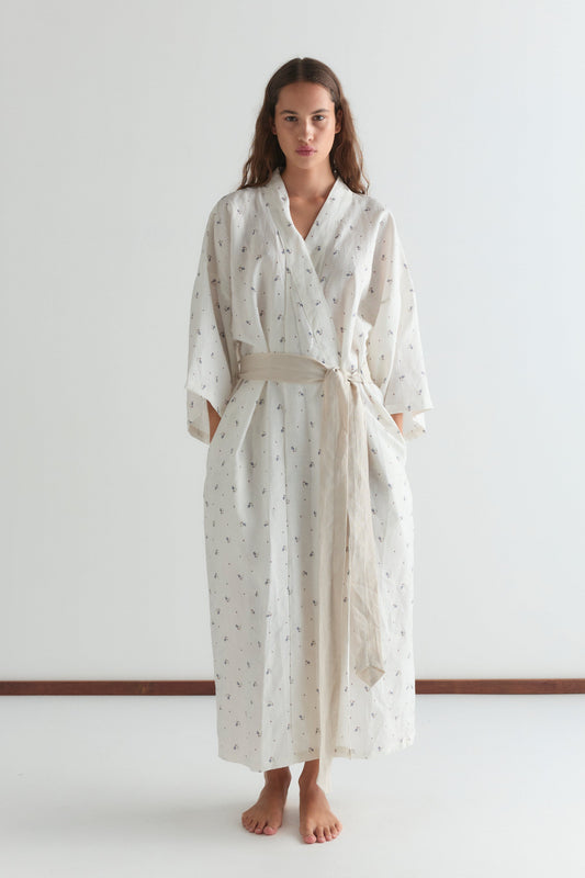 A woman stands barefoot against a white background, wearing the 02 robe - vintage floral. The full-length robe features a subtle pattern, linen waist tie, and pockets, adding a touch of effortless elegance to her look.