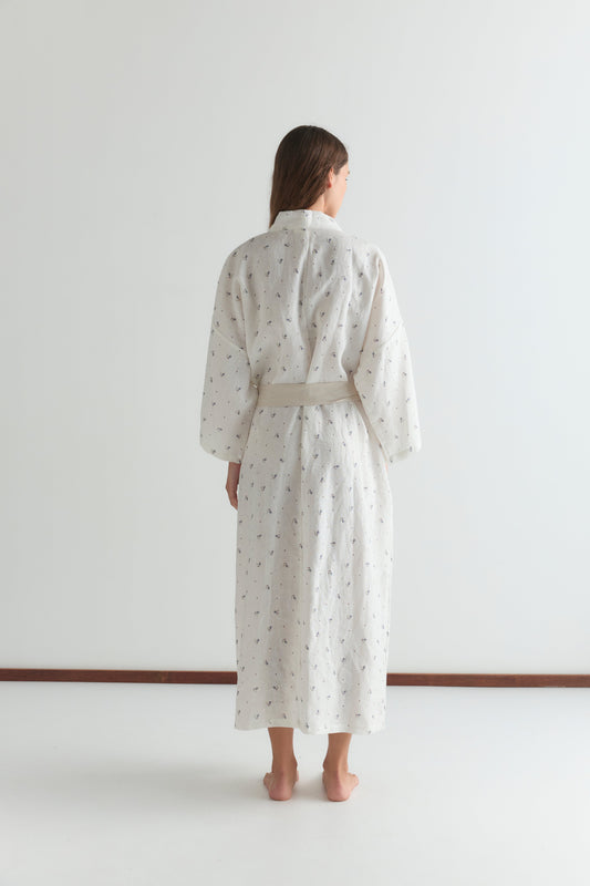 A woman stands barefoot, facing away, wearing the 02 robe - vintage floral with a linen waist tie and subtle print, set against a plain white background.