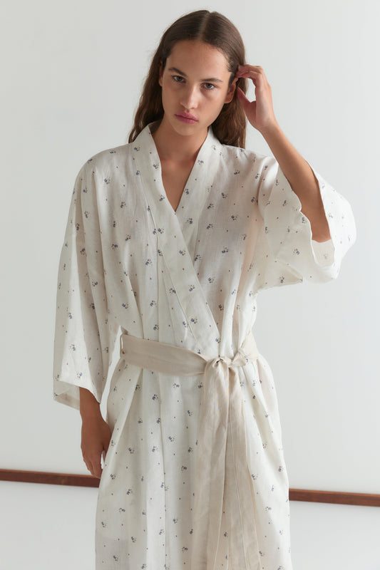 A woman with long brown hair models the 02 robe - vintage floral, featuring a linen waist tie. She stands against a white background, looks at the camera, and touches her hair with one hand.