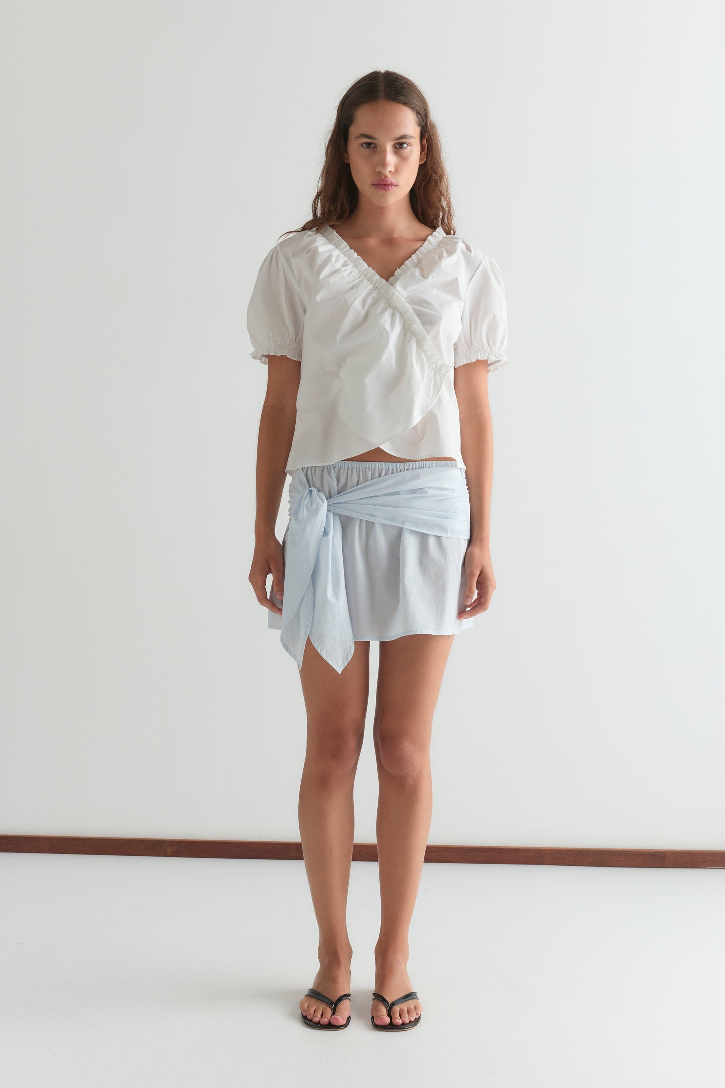 A woman with long brown hair and a neutral expression stands against a white wall wearing the Cross Frill Top in white, a light blue wrap skirt knotted at the waist, and black sandals.