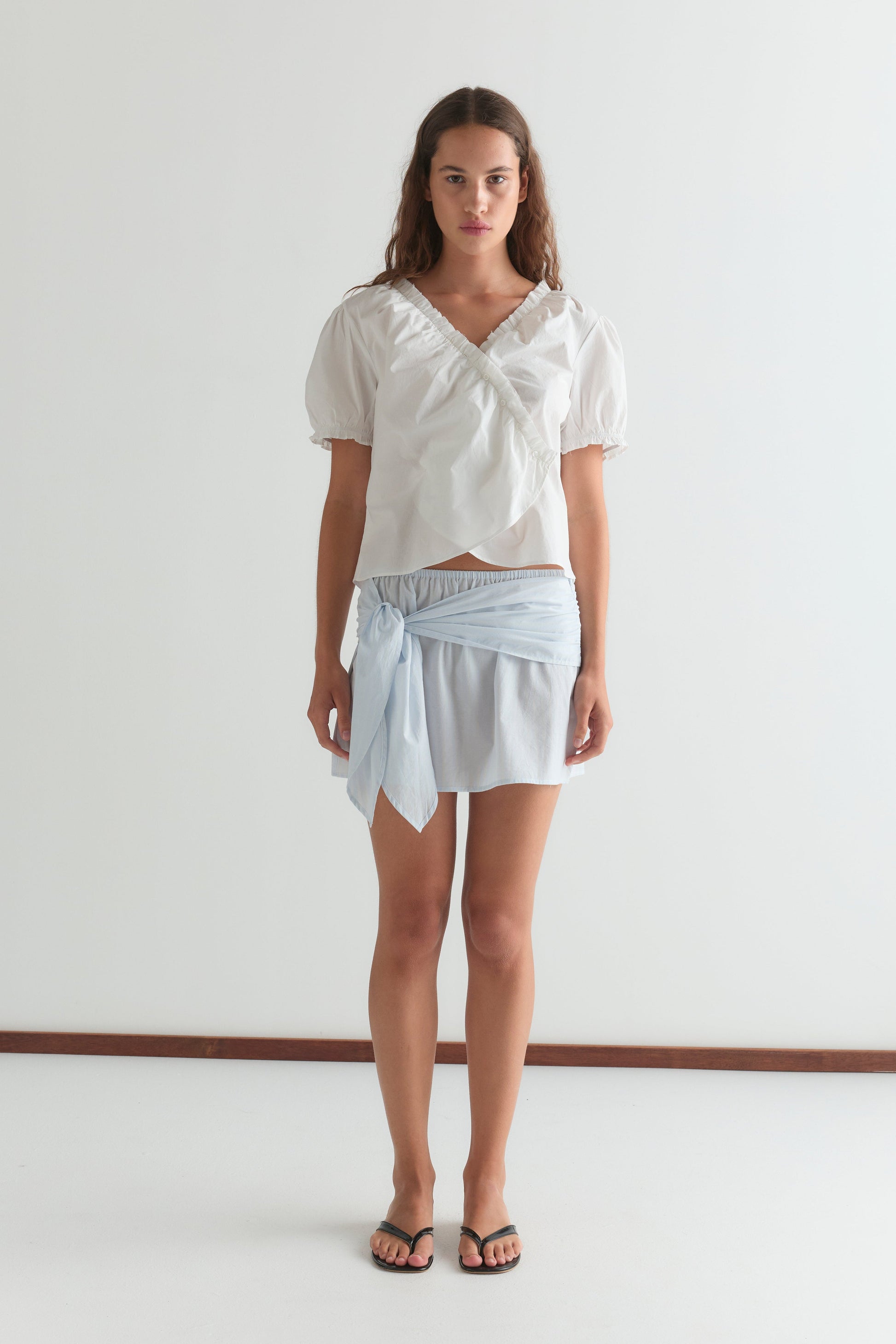 A woman with long brown hair and a neutral expression stands against a white wall wearing the Cross Frill Top in white, a light blue wrap skirt knotted at the waist, and black sandals.