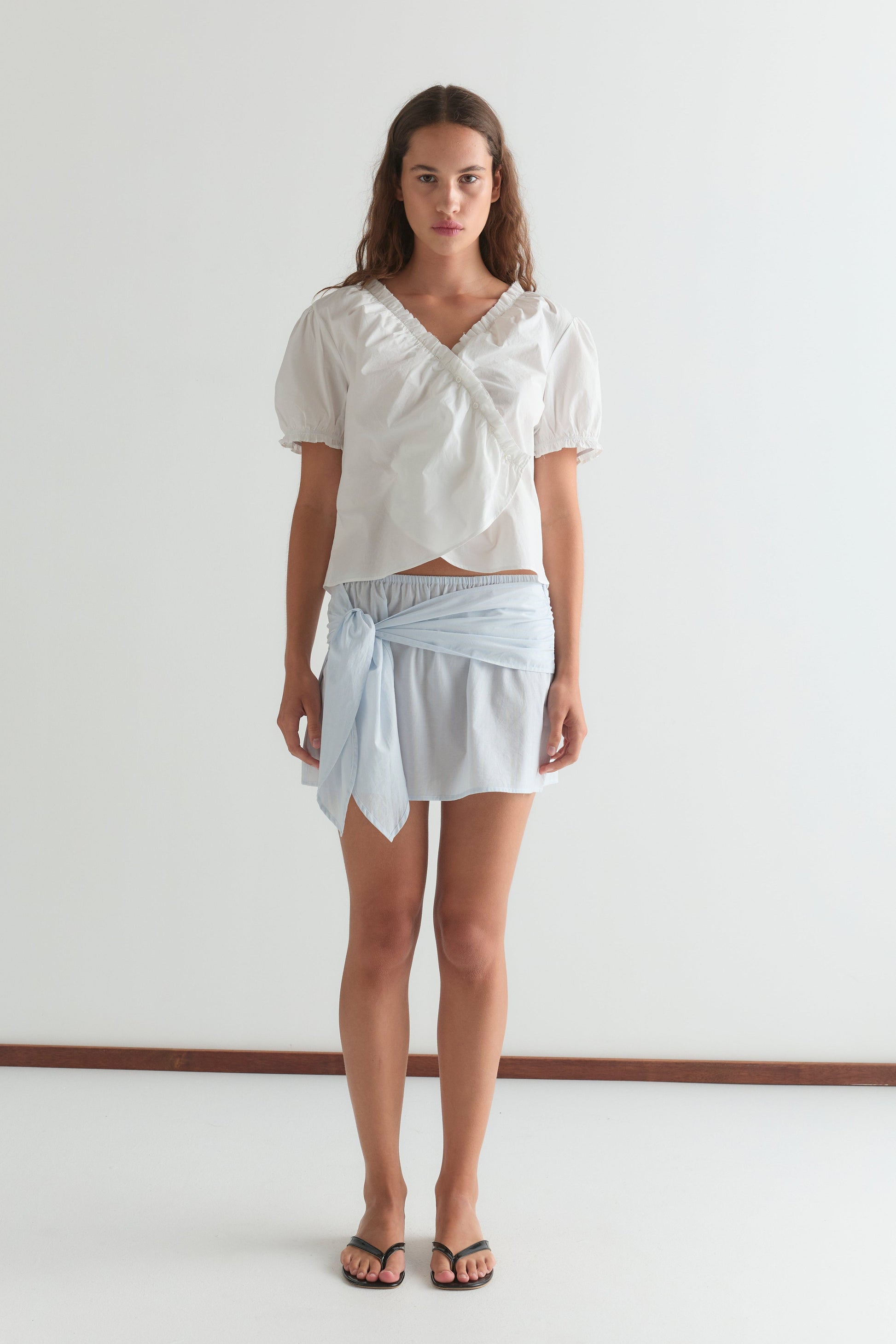 A woman with long brown hair wears a white puff-sleeve blouse, the Belted Skirt - Powder Blue Stripe from Deiji Studios, and black sandals while standing against a plain white wall with a neutral expression.