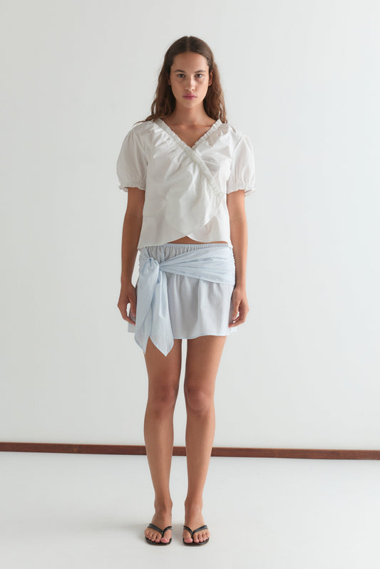 A woman with long brown hair wears a white puff-sleeve blouse, the Belted Skirt - Powder Blue Stripe from Deiji Studios, and black sandals while standing against a plain white wall with a neutral expression.