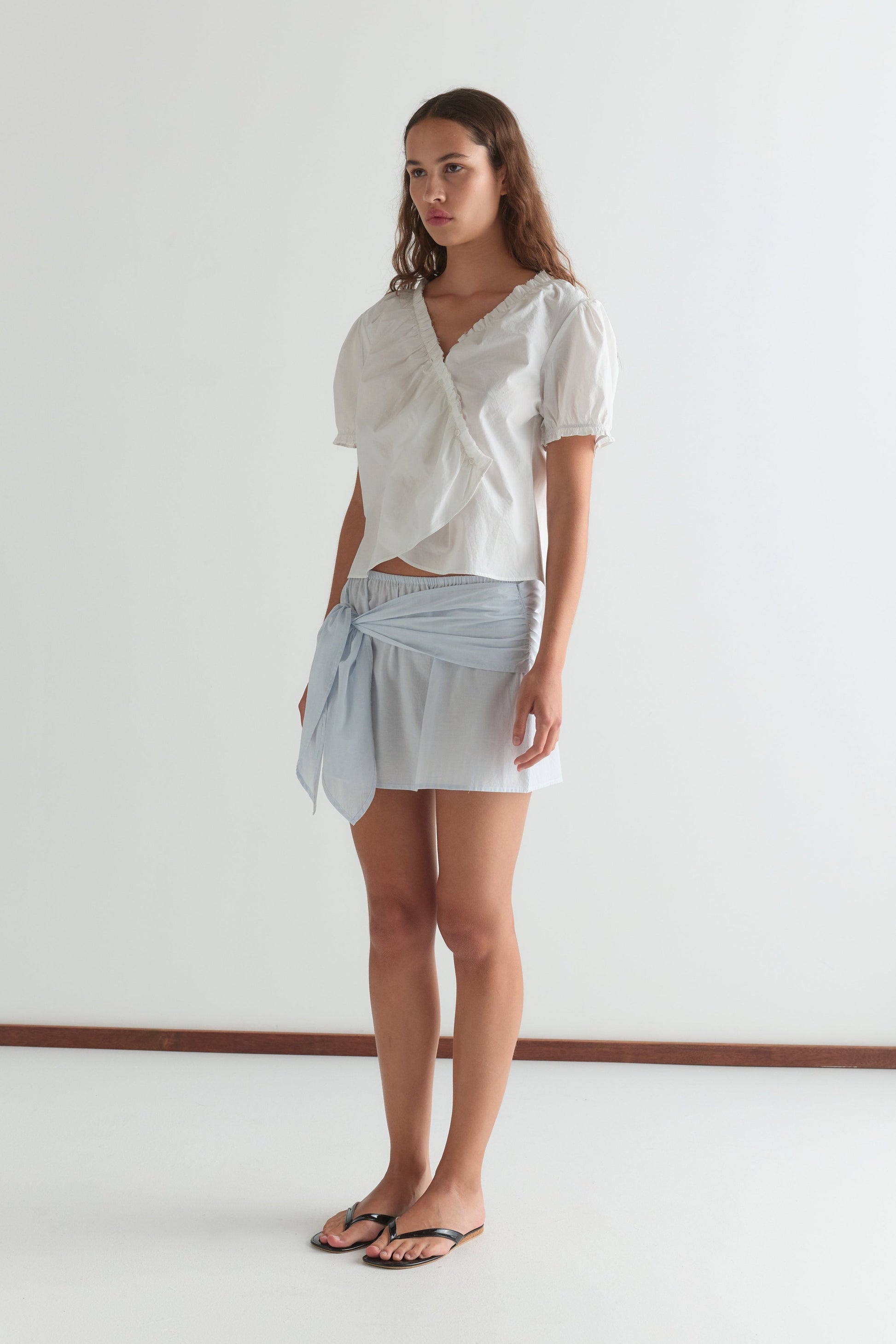 A woman stands indoors in a Cross Frill Top - White, paired with a light blue tie mini skirt and black flip-flops. She has long brown hair and a neutral expression, set against a plain light background.