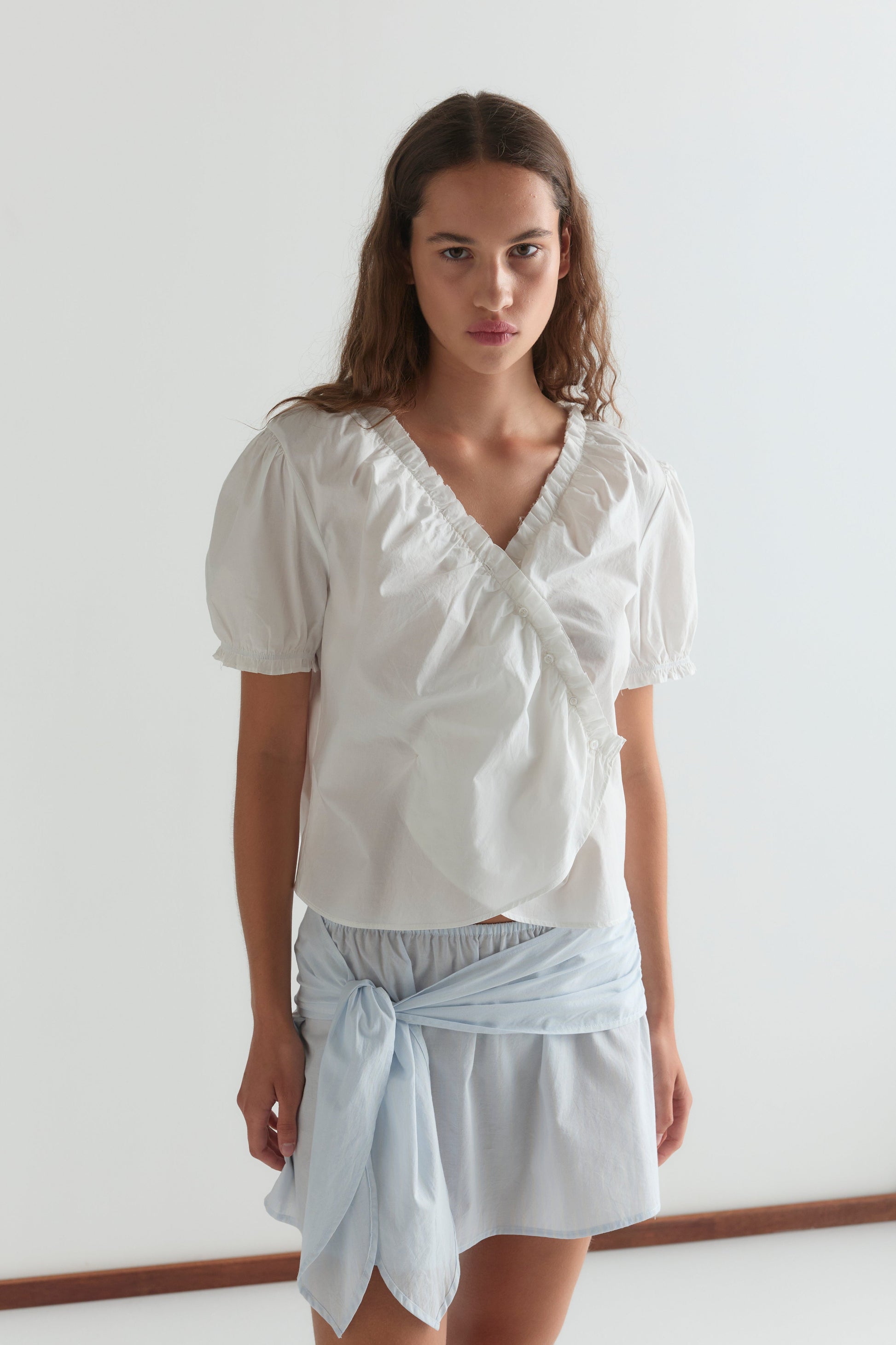 A woman stands against a plain white background wearing the Cross Frill Top, a white short-sleeved wrap blouse, paired with a light blue skirt featuring a matching sash tied at the waist.