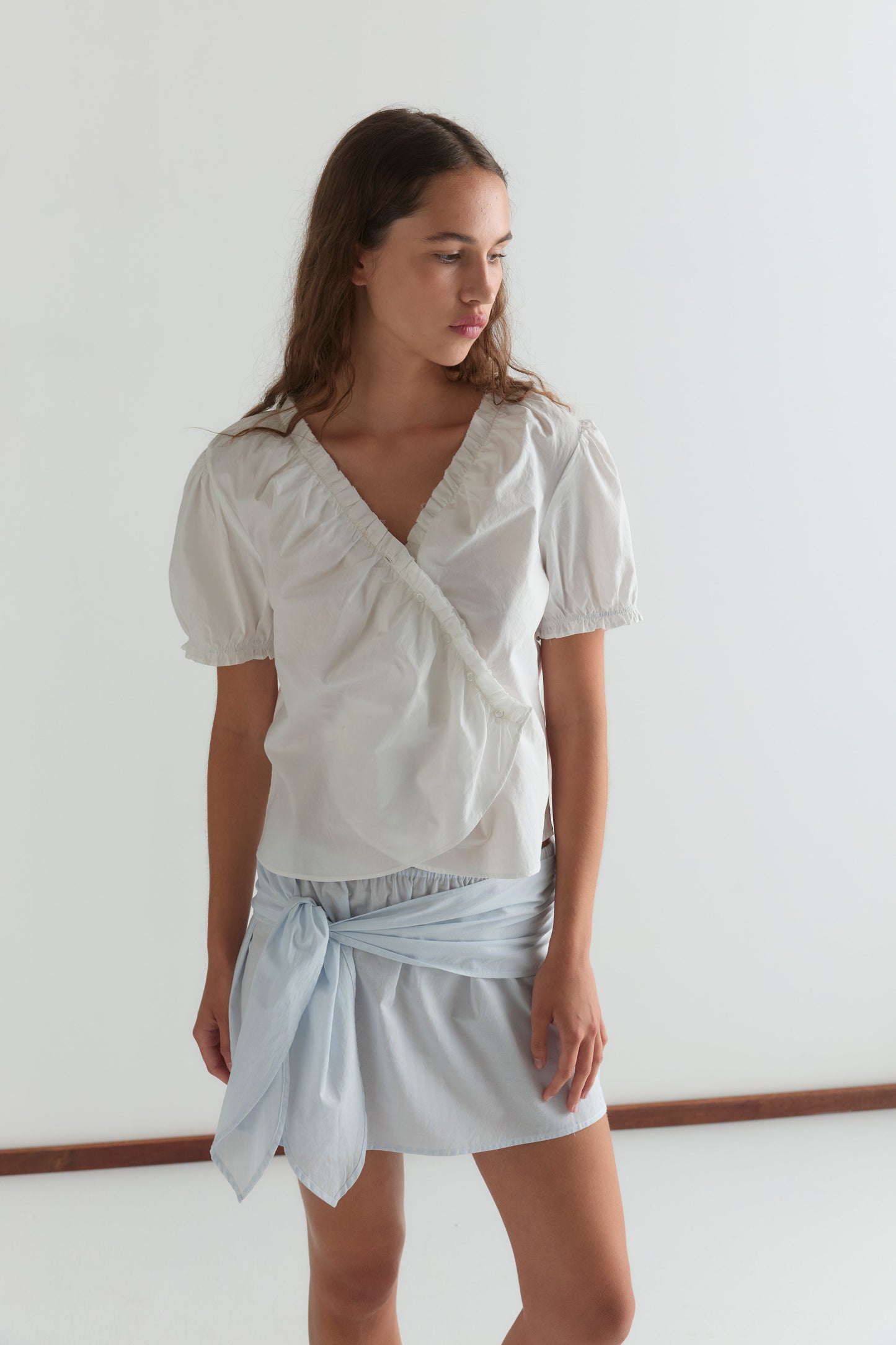 A young woman with long brown hair wears the Cross Frill Top in white, featuring a cropped relaxed fit, paired with a light blue skirt with a fabric tie detail, standing against a plain white background and looking down to her left.