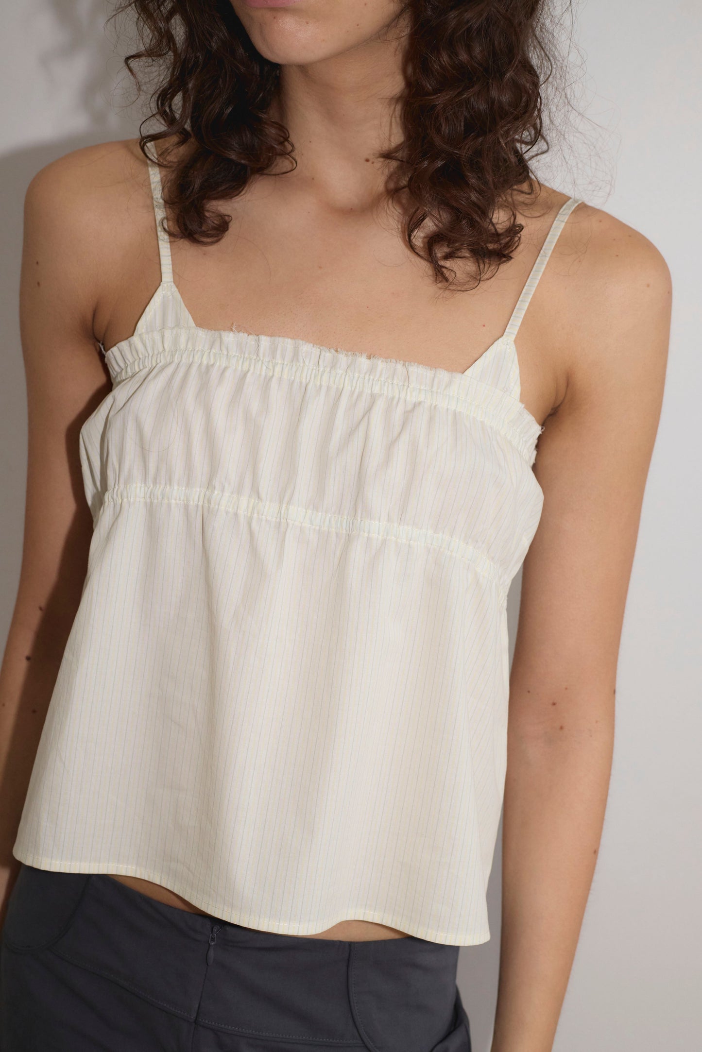 A woman with wavy brown hair wears the Tri Cami - Citrus Stripe by Deiji Studios, a lightweight white top with subtle vertical stripes, paired with dark pants. She stands against a plain white background, her face partially out of frame.