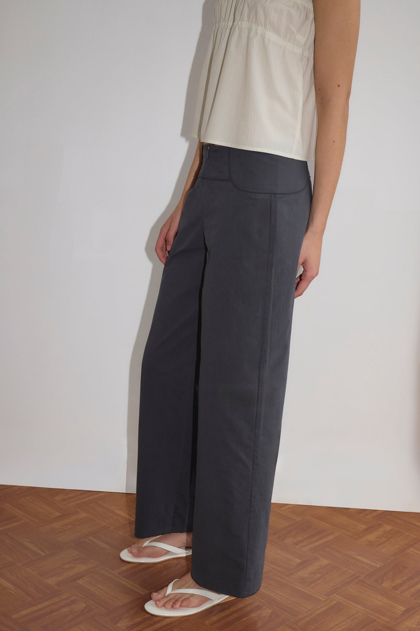 A person stands on a wooden floor wearing Deiji Studios' Panelled Pant - Charcoal, styled with a sleeveless white top and white flip-flops. The side view showcases the pant's relaxed fit and minimalist design.