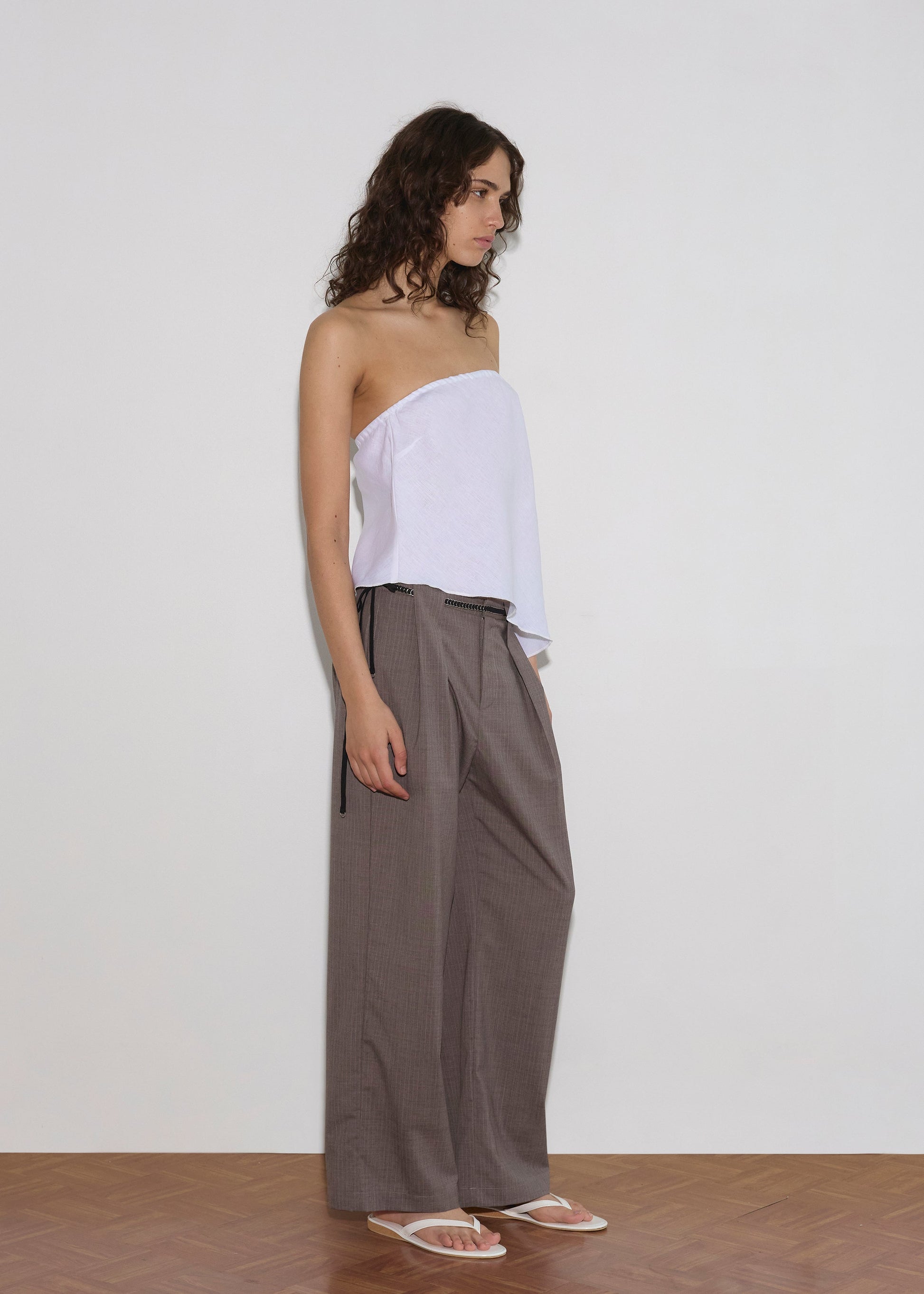 A person with curly hair stands against a white wall wearing the Line Top, loose gray wide-leg pants, and white sandals. The floor is wooden, and they are looking to the side.