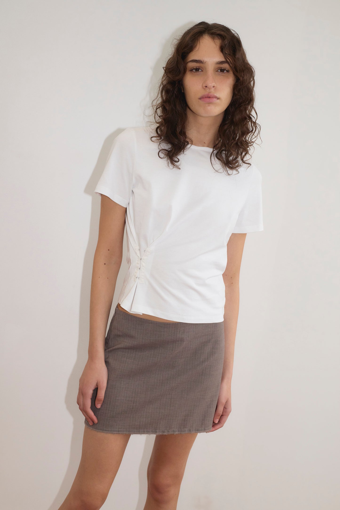 A person with long curly brown hair wears a white short-sleeve ruched top and the Eye Skirt - Grey Pink Stripe from Deiji Studios, standing against a plain white wall and looking at the camera with a neutral expression.