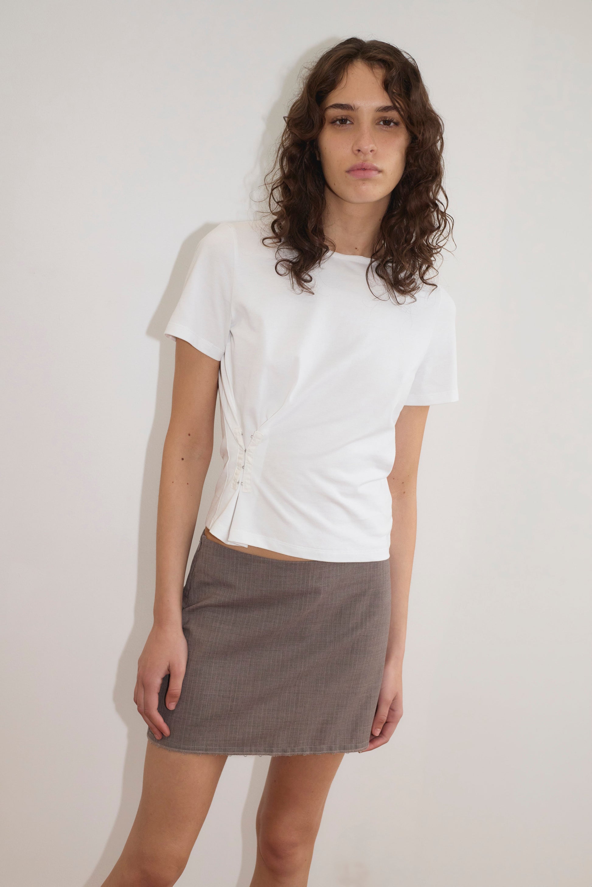 A person with long curly brown hair wears a white short-sleeve ruched top and the Eye Skirt - Grey Pink Stripe from Deiji Studios, standing against a plain white wall and looking at the camera with a neutral expression.