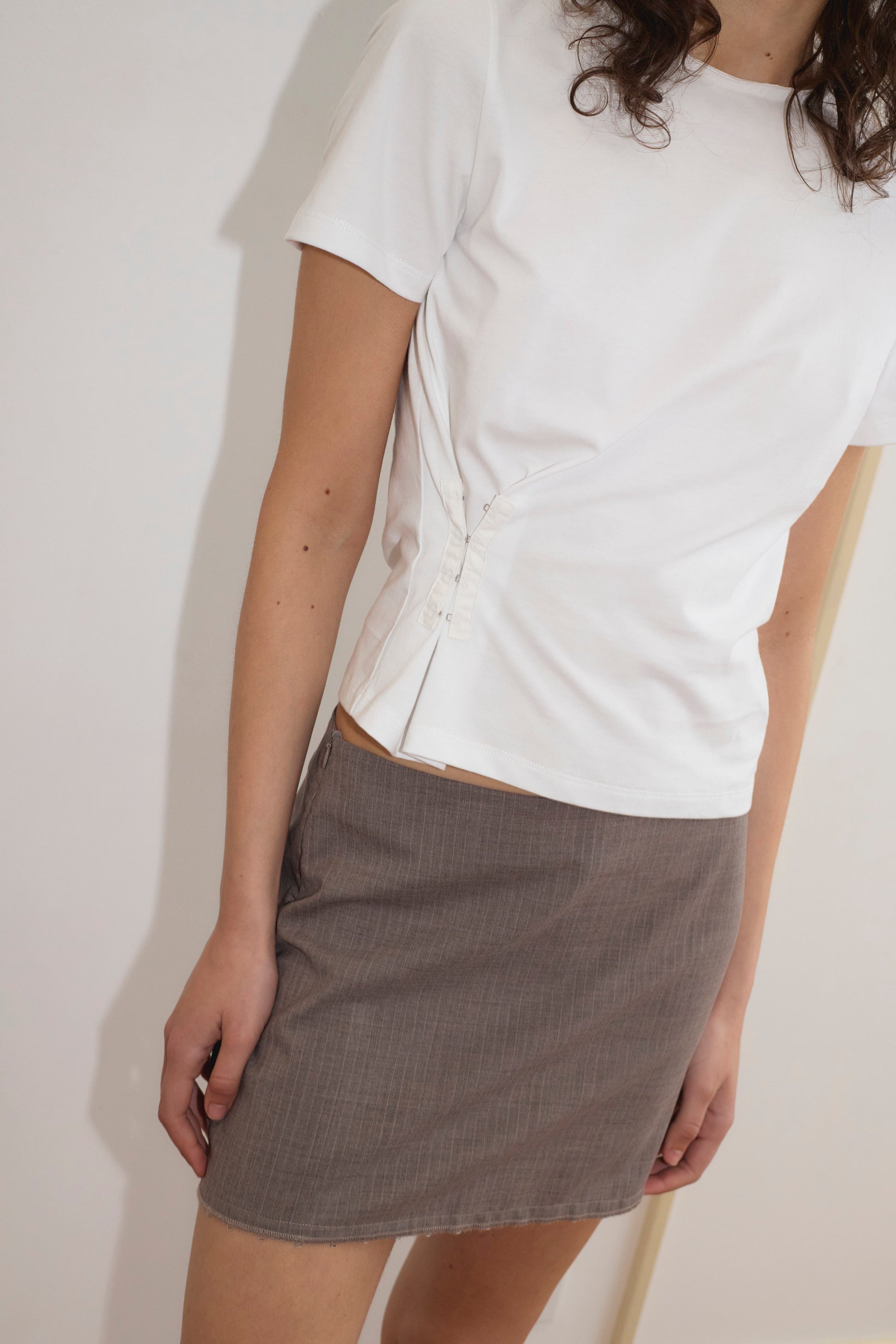 A person stands against a light background wearing a white short-sleeve top with side button details and Deiji Studios' Eye Skirt in Grey Pink Stripe. Their head is partially out of frame.