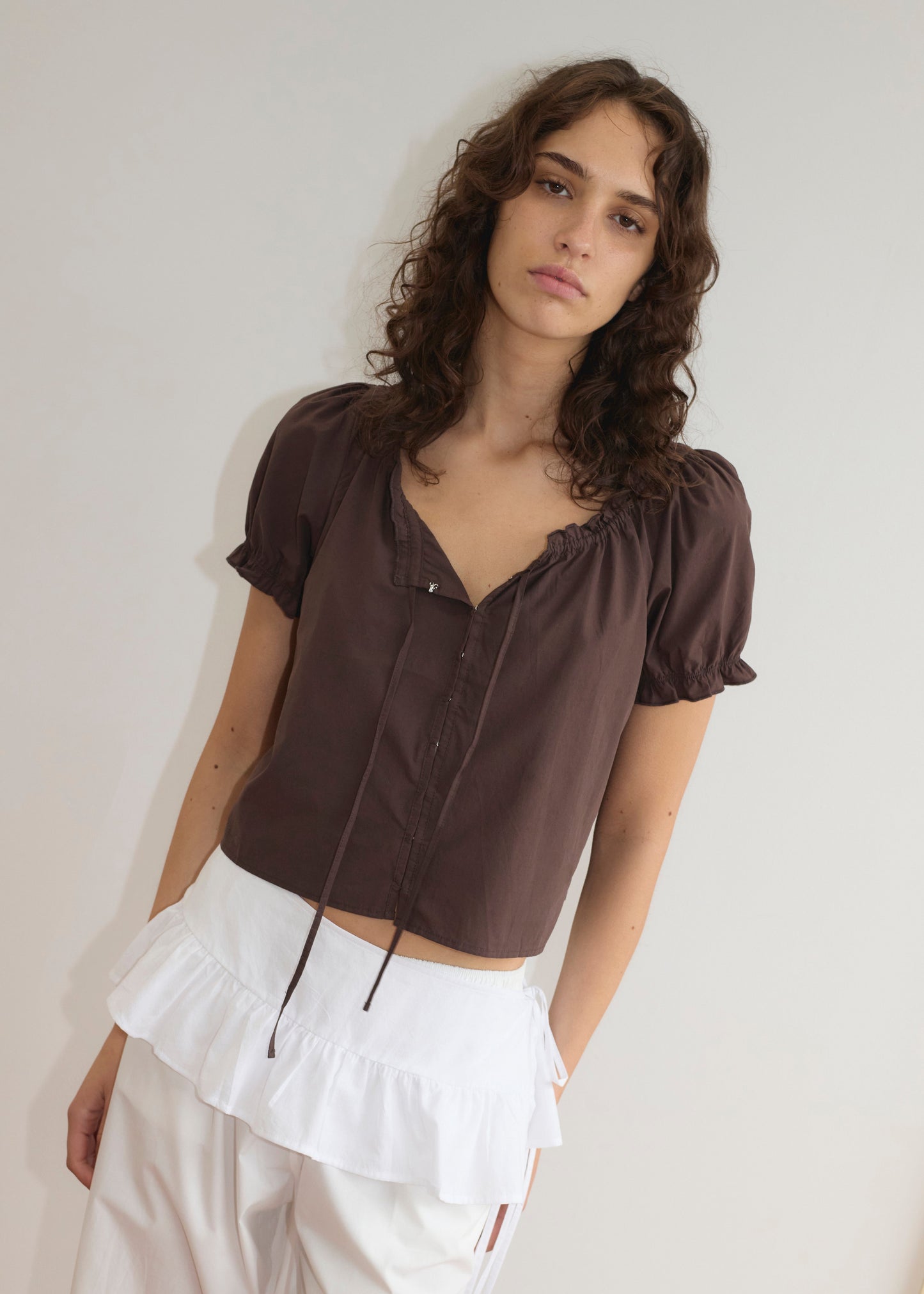 A young woman with wavy brown hair wears a brown short-sleeve blouse and the Flounce Belt - White, which adds a lightweight frill ruffle detail to her pants. She stands against a plain off-white background, looking at the camera.